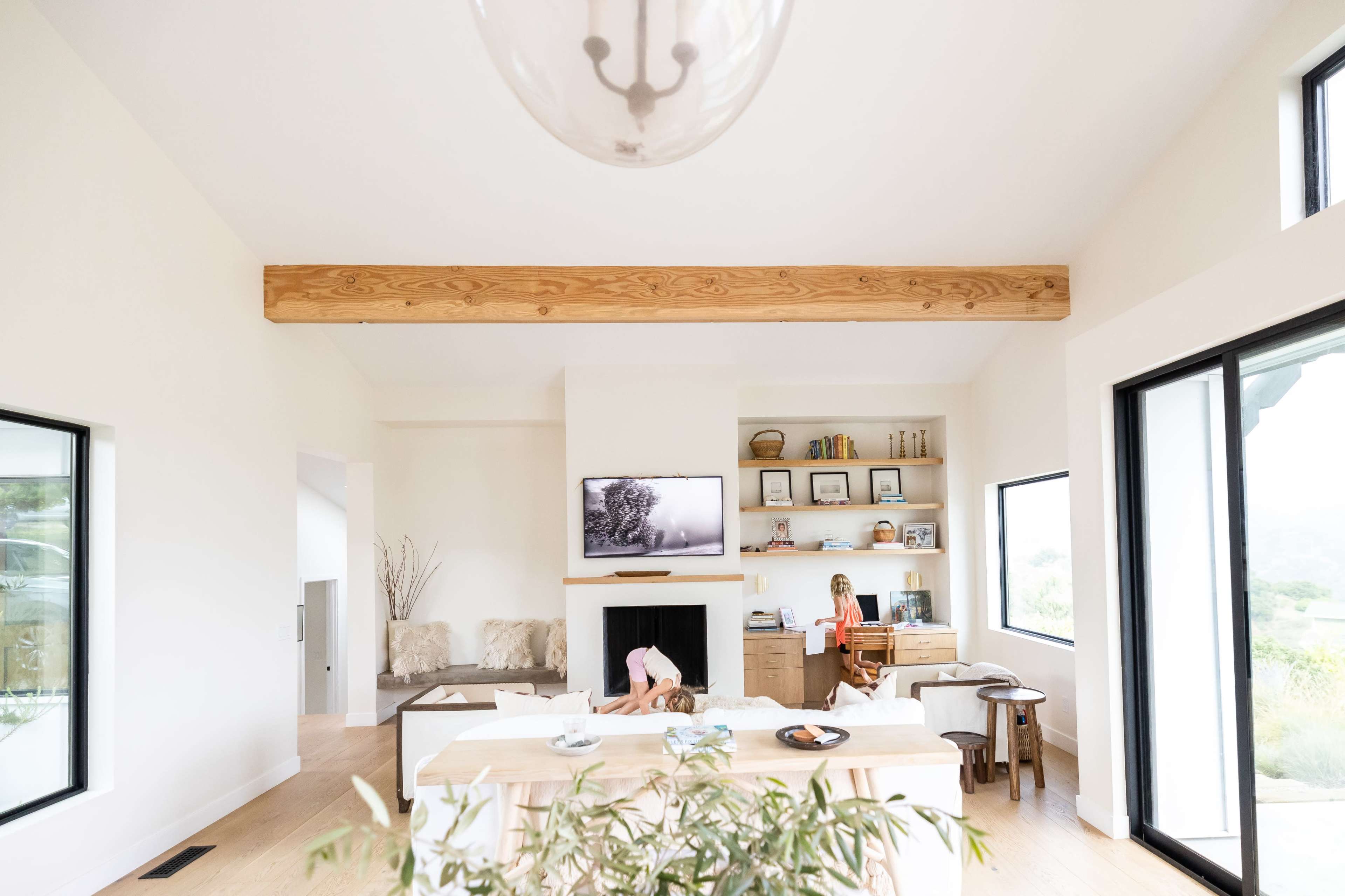 A bright, modern living room features a large wooden beam overhead, a television mounted on the wall, and a woman seated at a desk while a child plays on the floor.