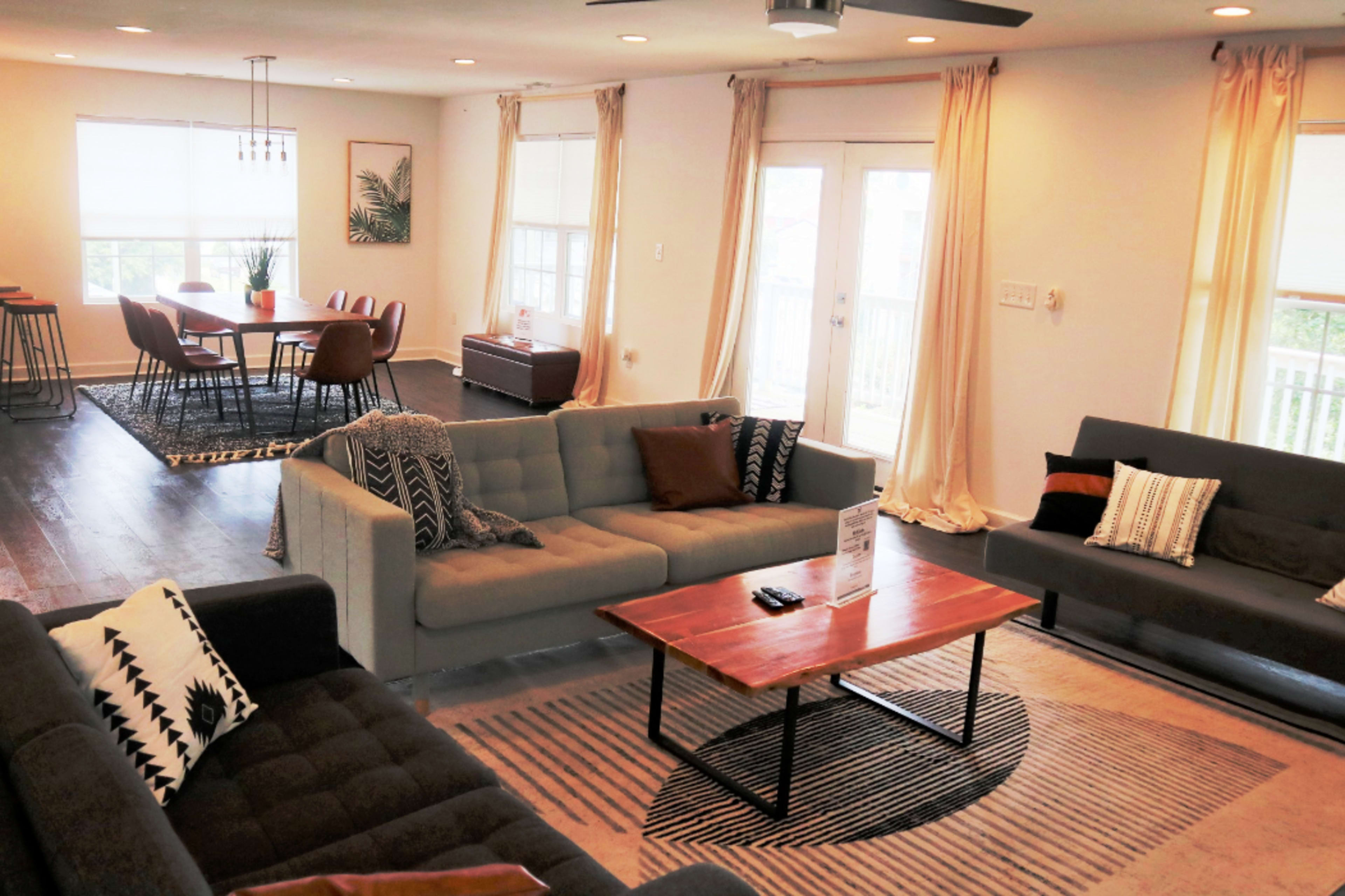 The living area features two sofas, a coffee table, and a dining table with chairs, all arranged in a bright, open space with large windows and wooden flooring.