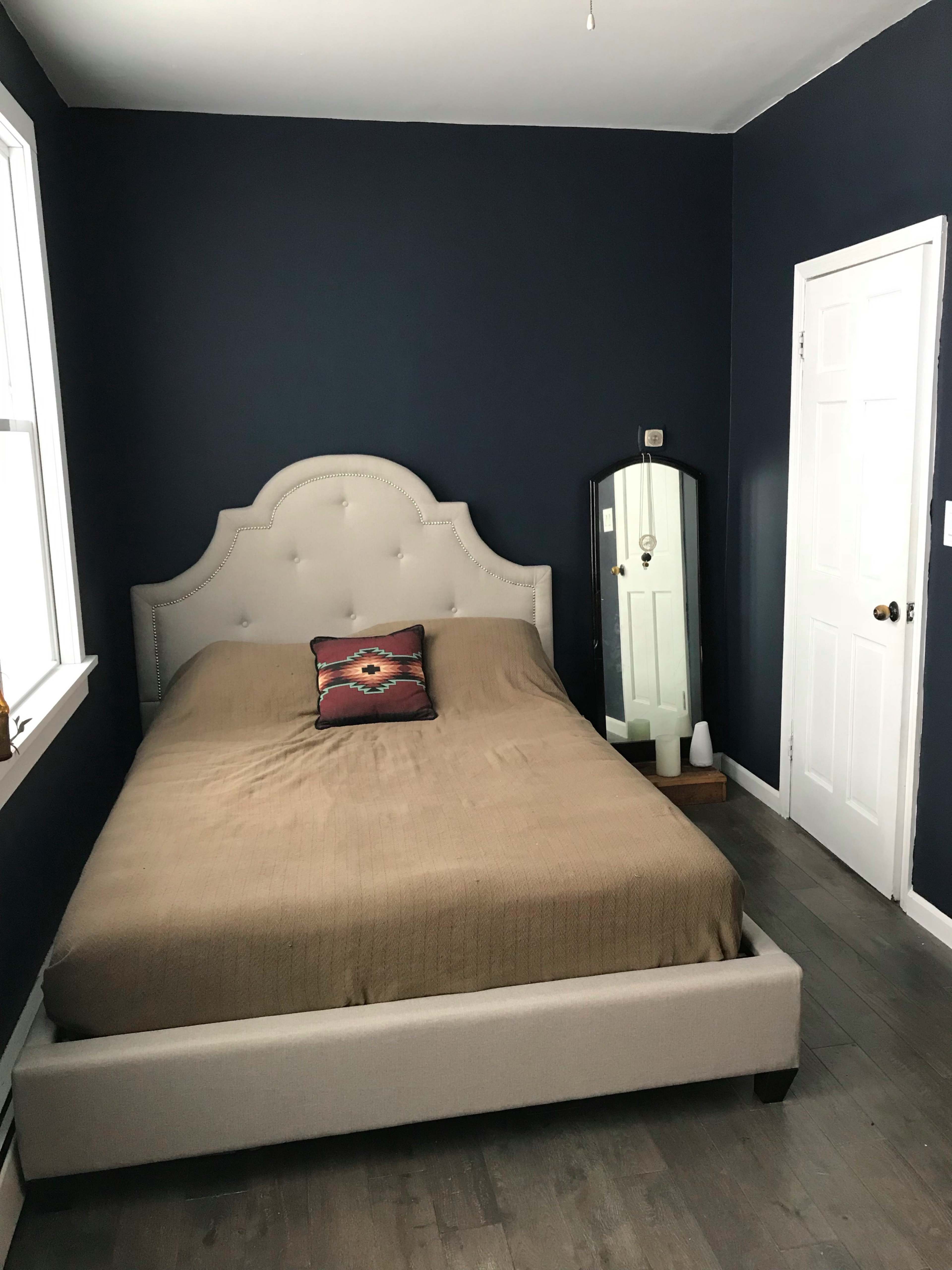 The image shows a simple bedroom with a bed featuring a tan coverlet, a patterned pillow, a full-length mirror, and dark blue walls.