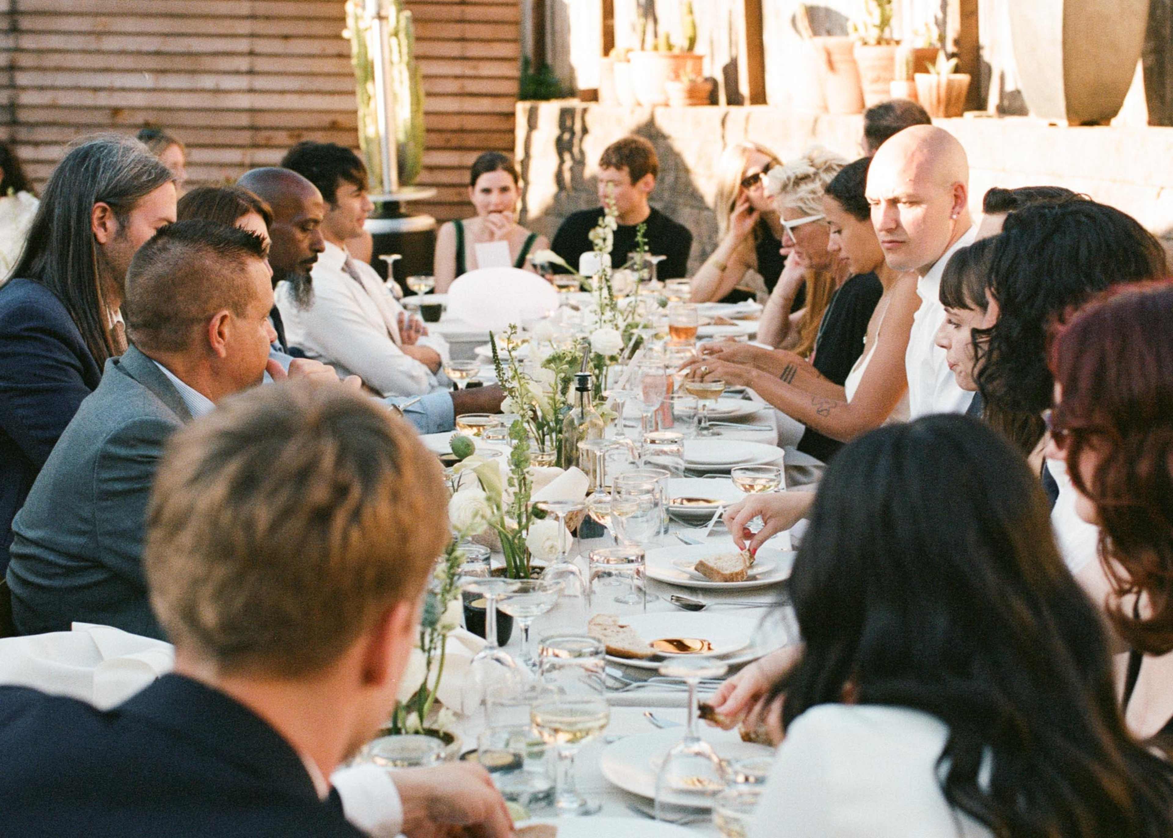 A long dining table is set outdoors, surrounded by a diverse group of people engaged in conversation while enjoying a meal.