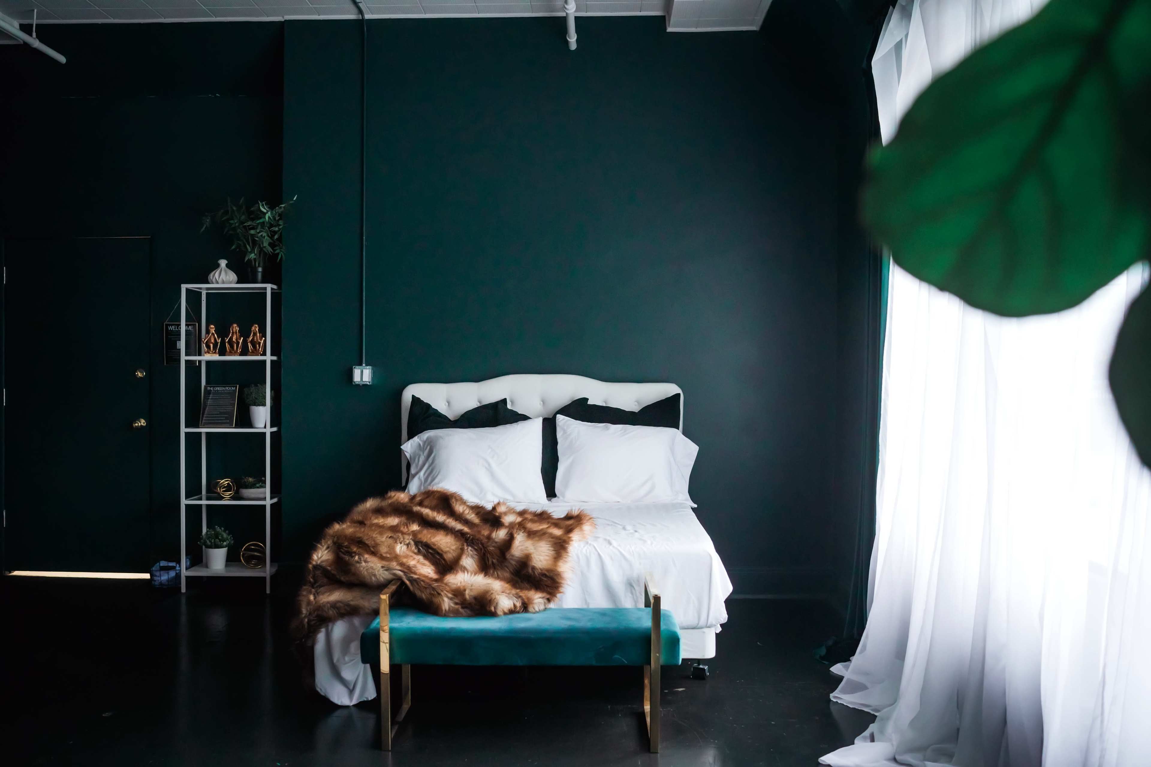 A cozy bedroom features a white bed with crisp linens, a fur throw blanket, and a teal bench in front, set against dark green walls.