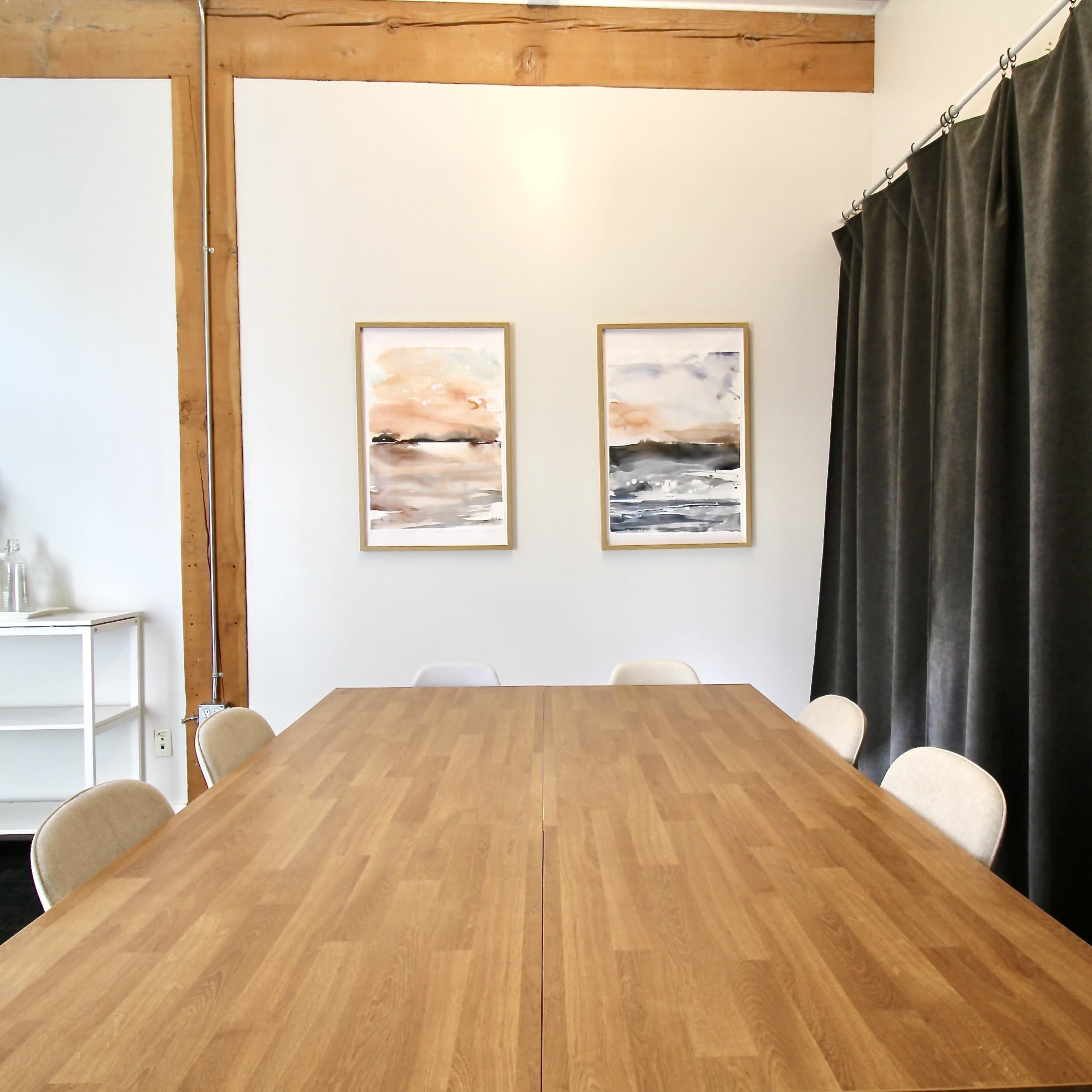 A wooden conference table is centered in a room with gray curtains and two framed paintings on the wall.