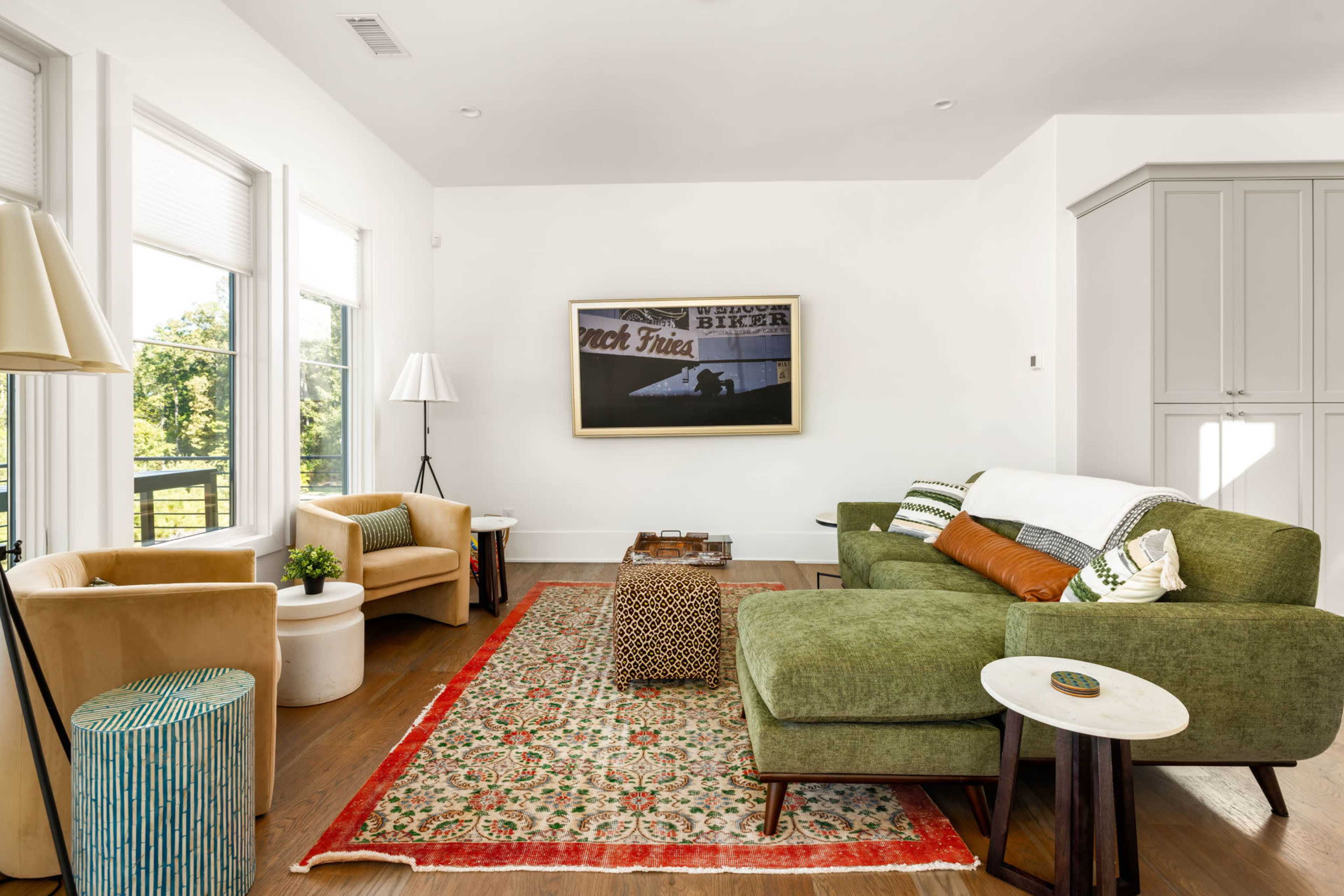 A modern living room features a green sofa, two beige armchairs, a patterned area rug, and a white side table against a backdrop of large windows.