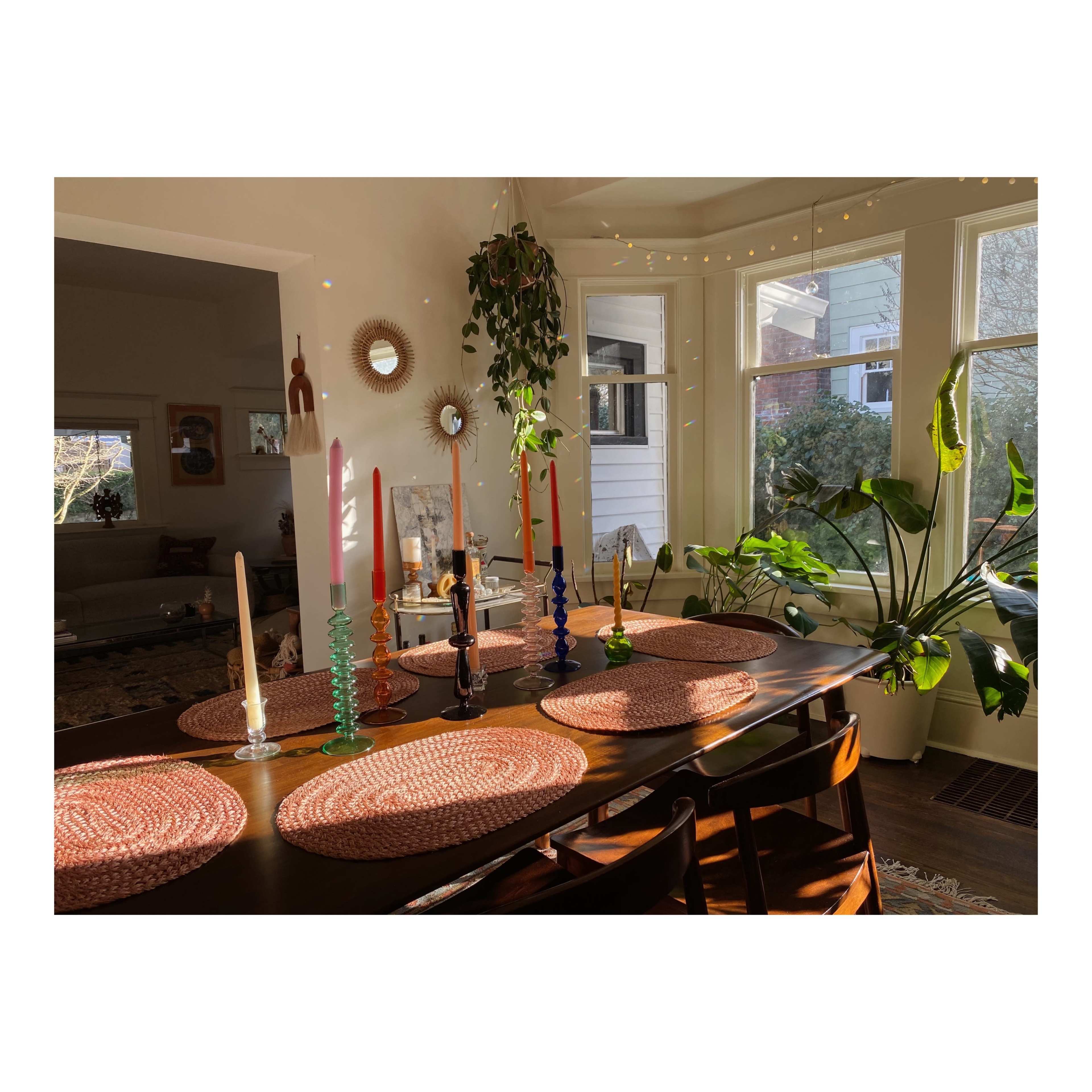 A dining table is set with colorful candles and woven placemats, illuminated by natural light from nearby windows.