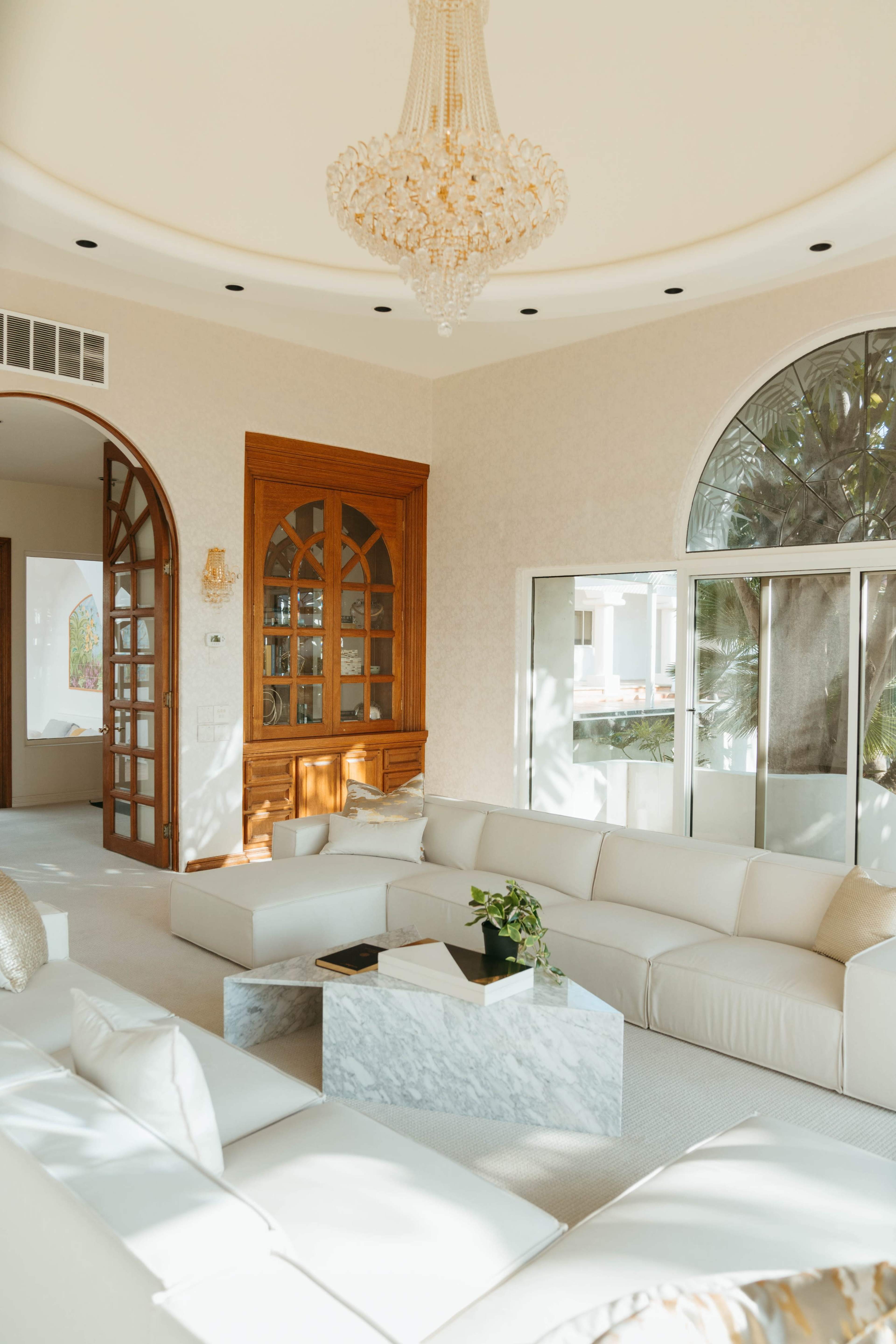 A spacious living room featuring a white sectional sofa, a marble coffee table, and a chandelier overhead, with large windows and a wooden cabinet in the background.