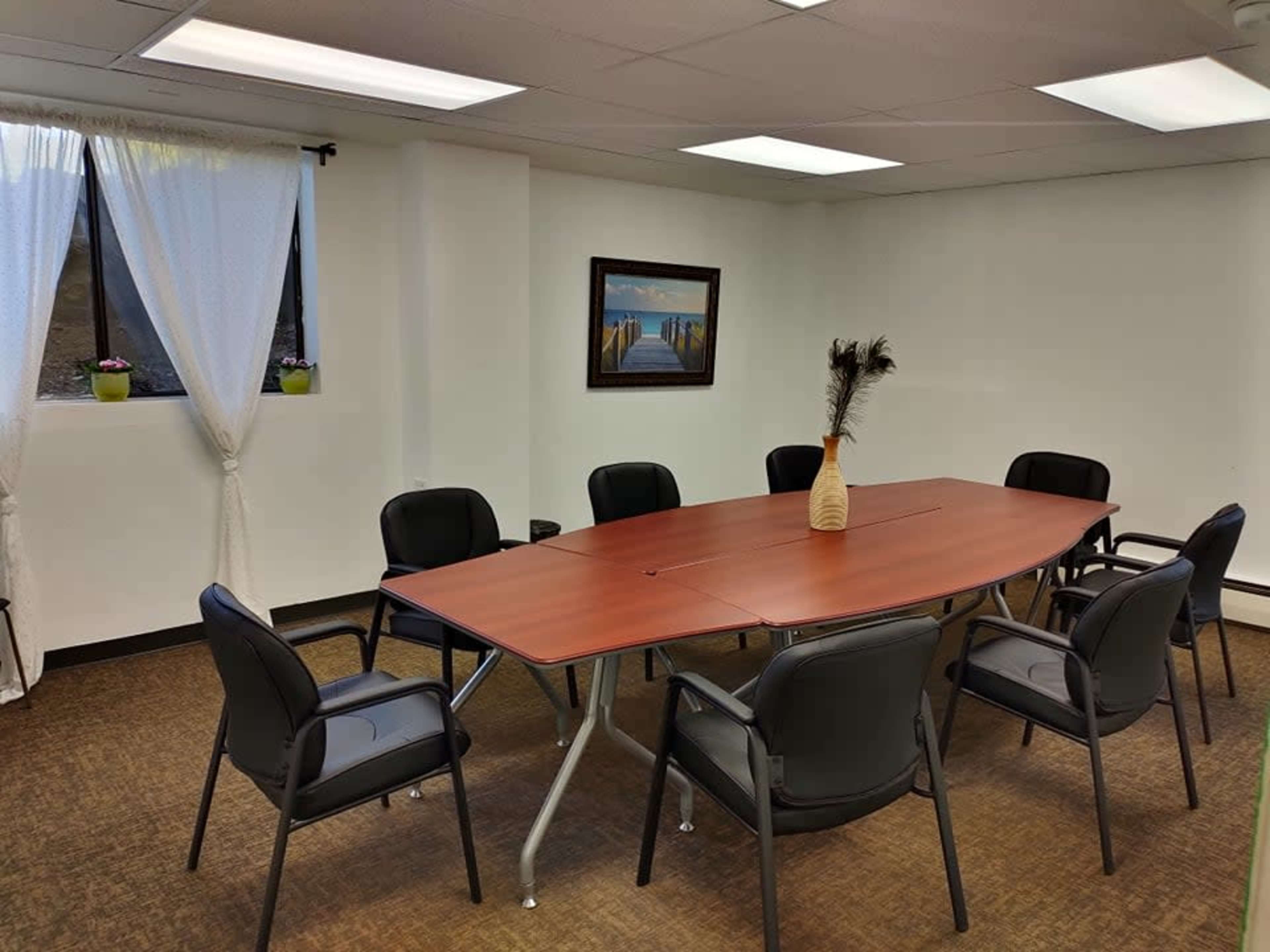 The image shows a conference room with an oval table surrounded by eight black chairs, and a painting on the wall across from a window with white curtains.