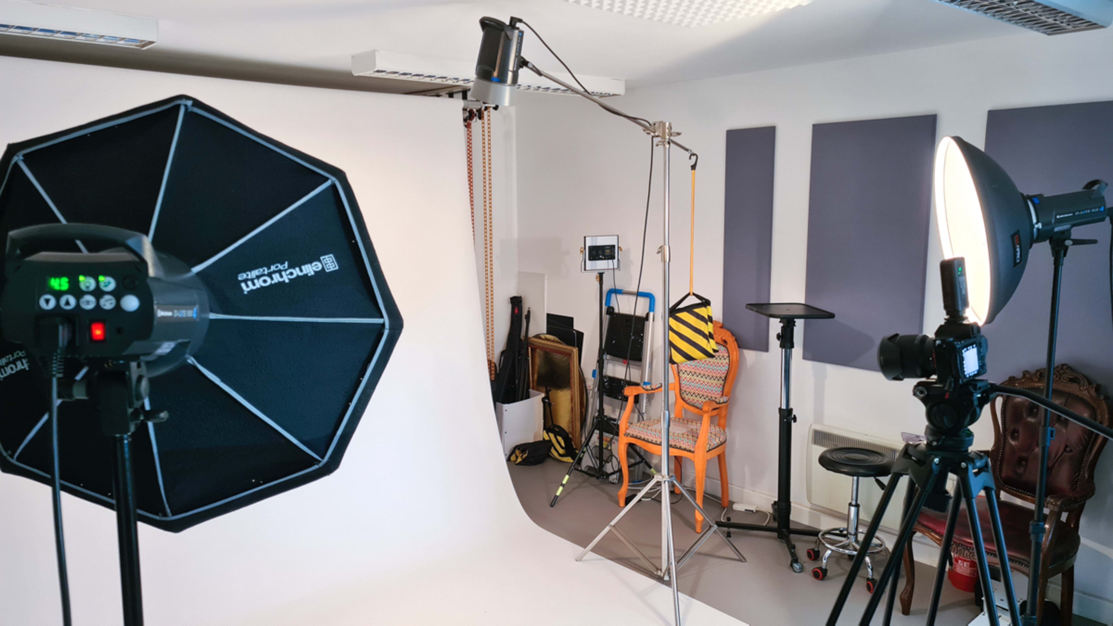 A photography studio setup with softbox lighting, a backdrop, and various support equipment arranged around a chair.