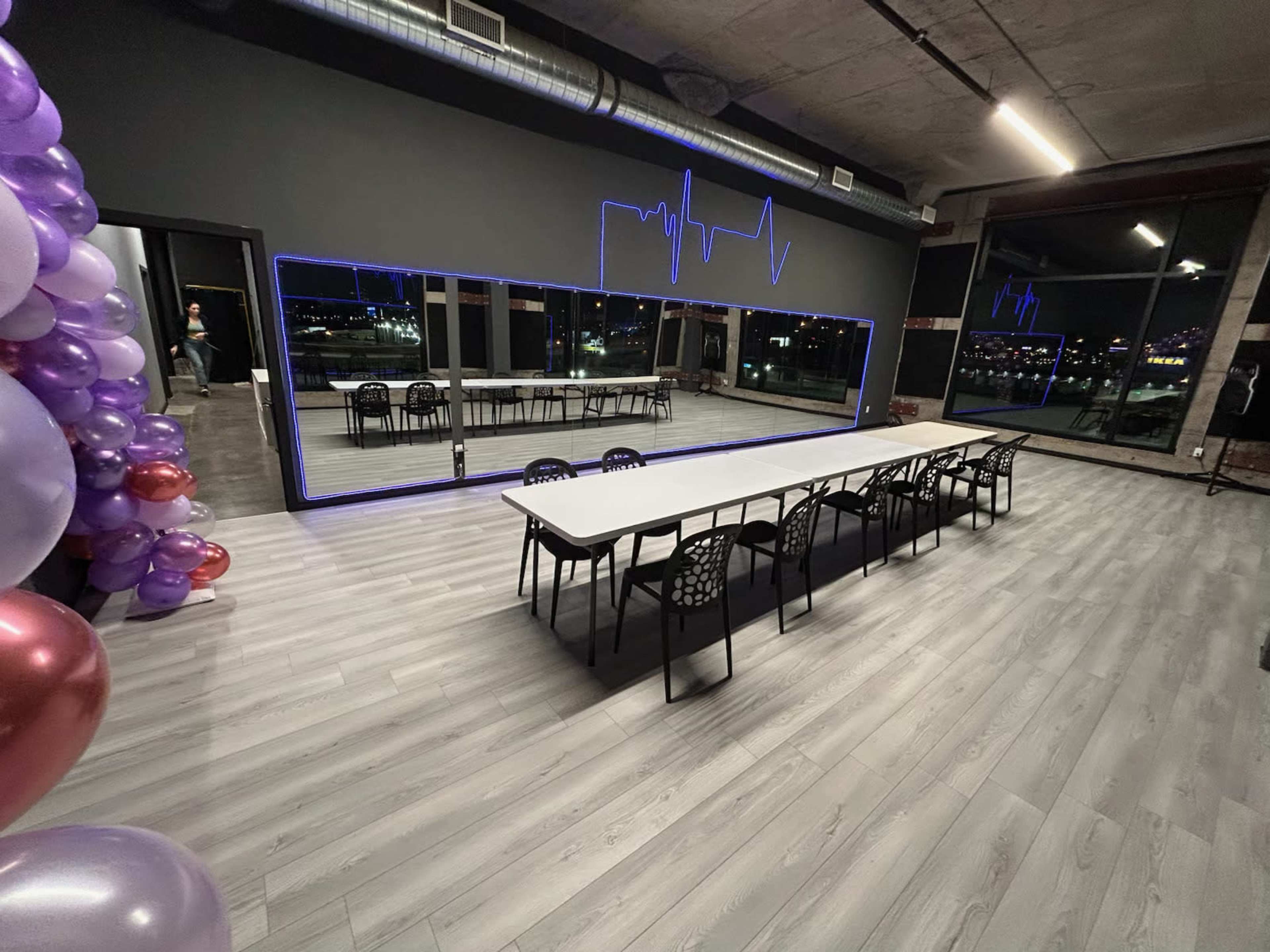 The image shows a modern event space with a long, white table surrounded by black chairs, a large illuminated mirror along one wall, and decorative balloons on the left side.
