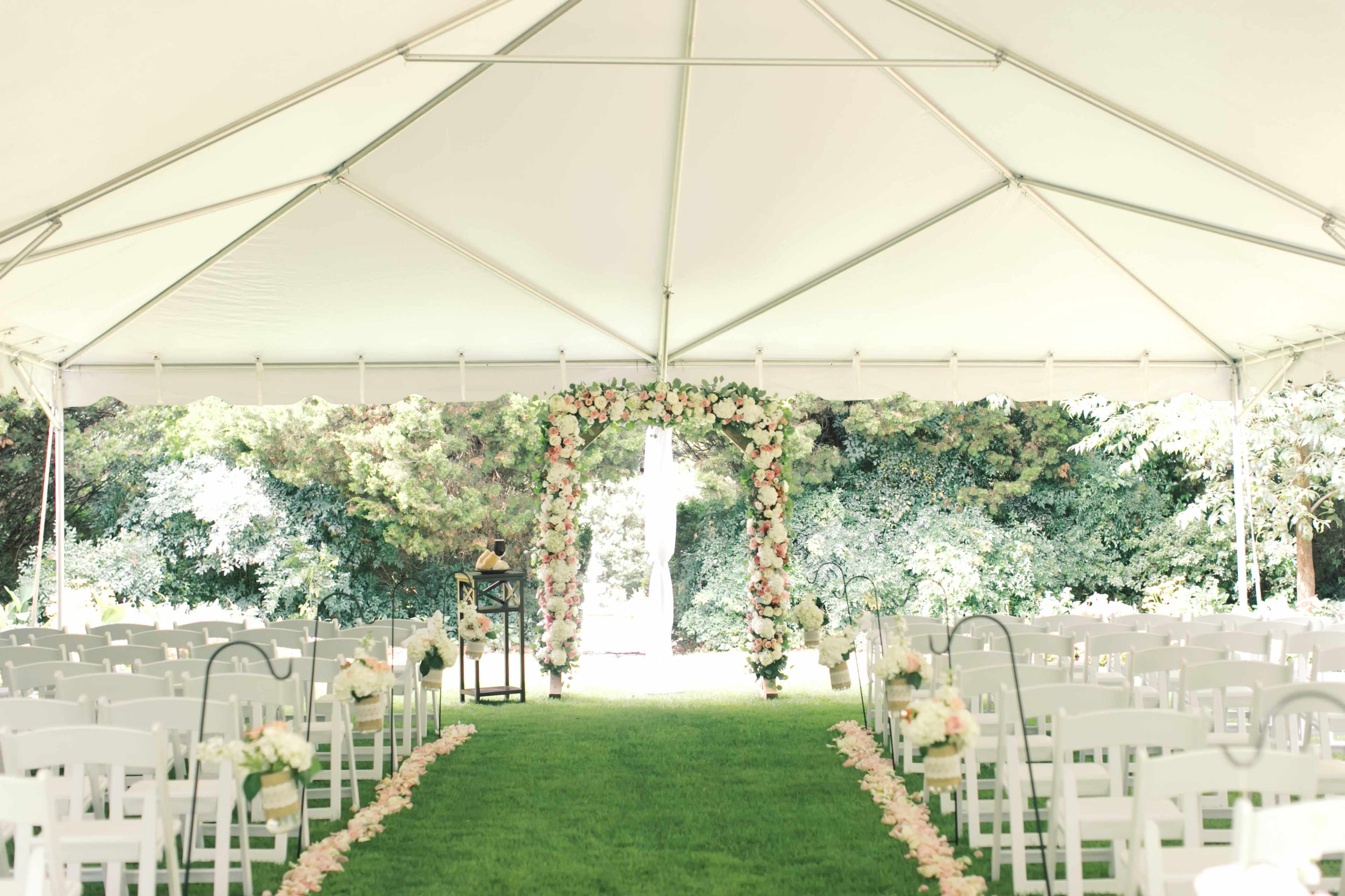 A decorated outdoor wedding ceremony under a tent features an arch adorned with flowers and a central aisle lined with floral arrangements and chairs.