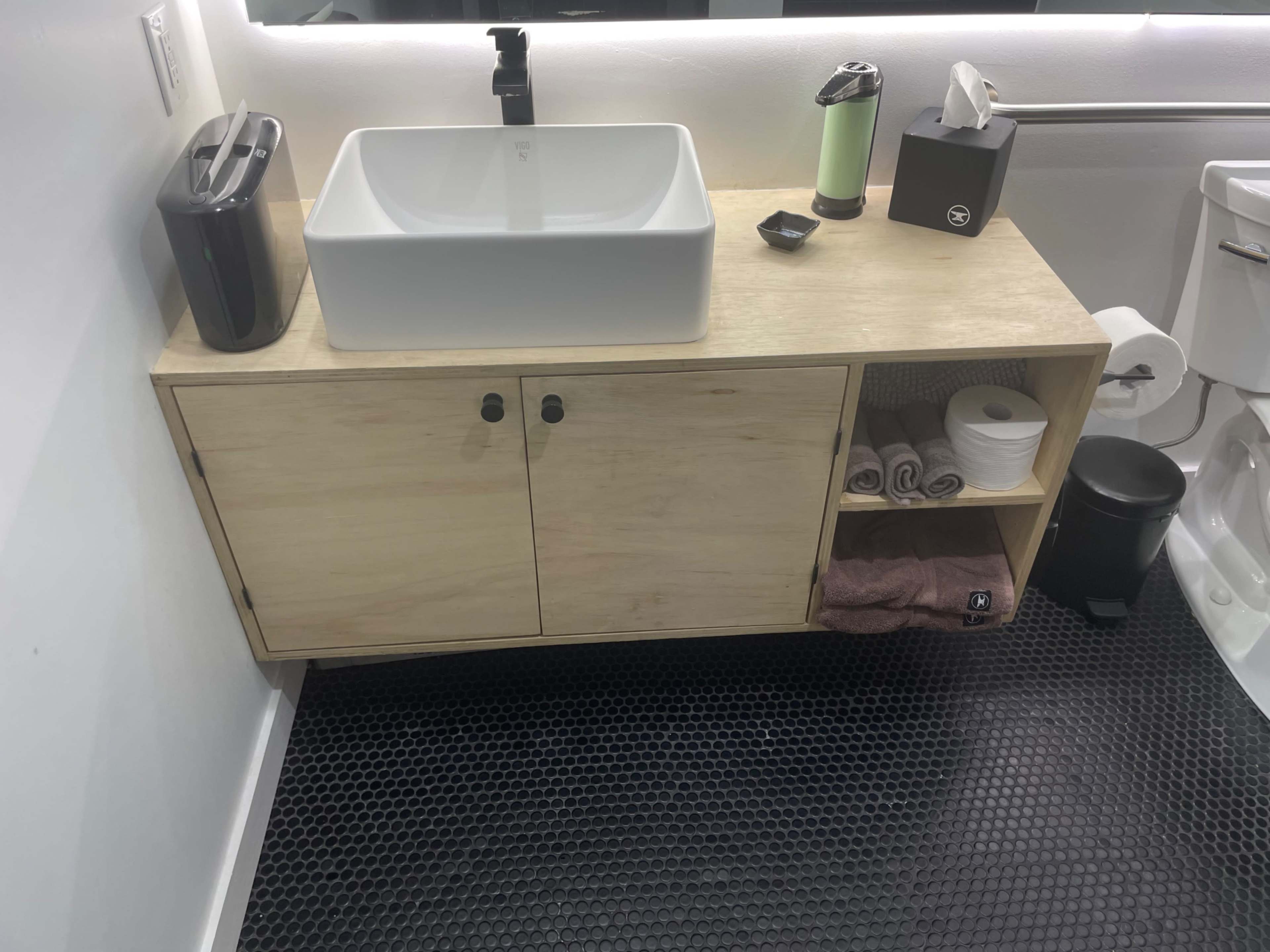 A modern bathroom sink area with a wooden cabinet, a white basin, a soap dispenser, towels, and a toilet in the background.
