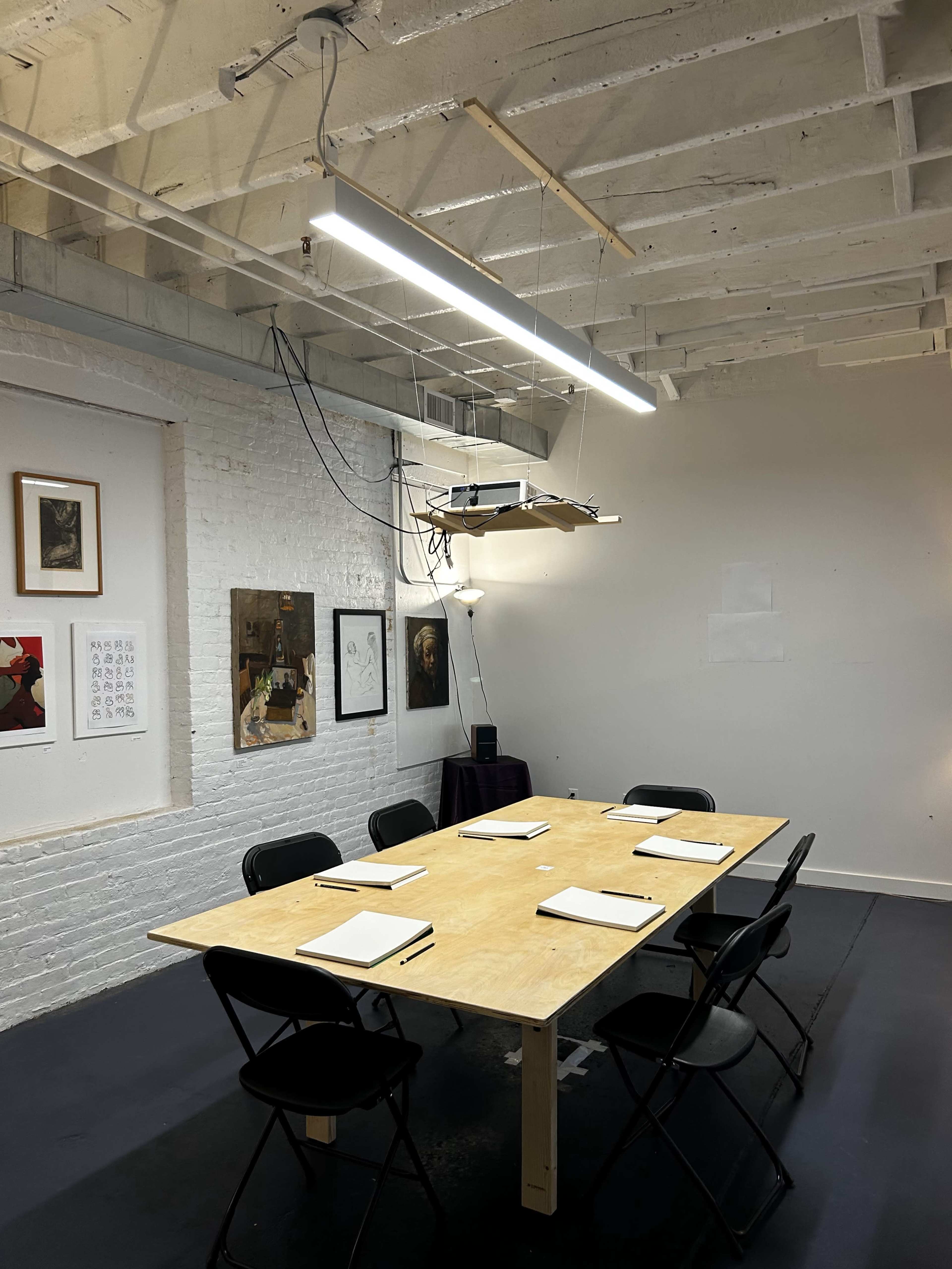 A spacious, minimalist meeting room features a wooden table surrounded by black chairs, with overhead lighting and artwork on the white brick walls.