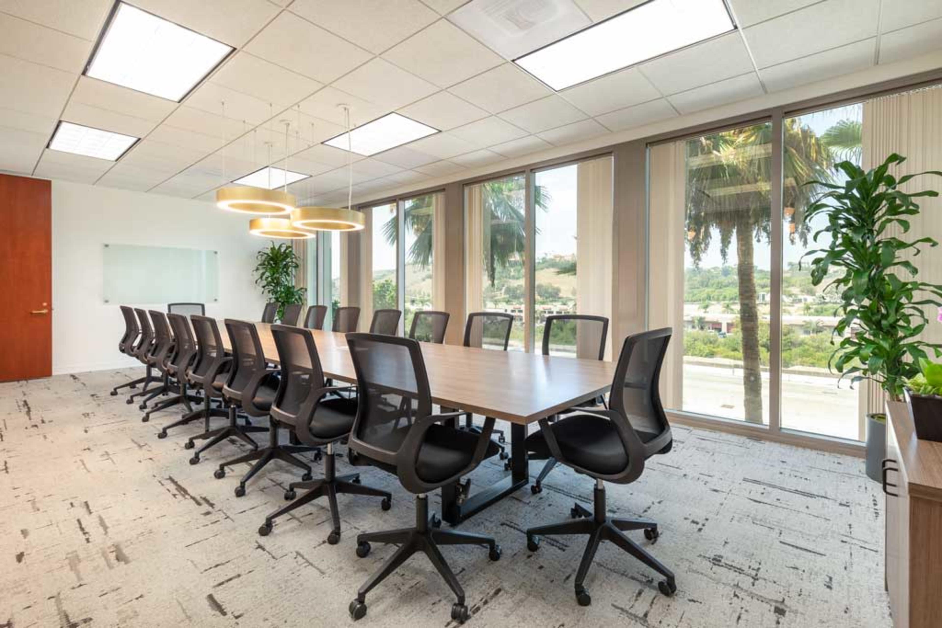 A modern conference room features a long wooden table surrounded by ergonomic chairs, with large windows that offer a view of palm trees and greenery outside.