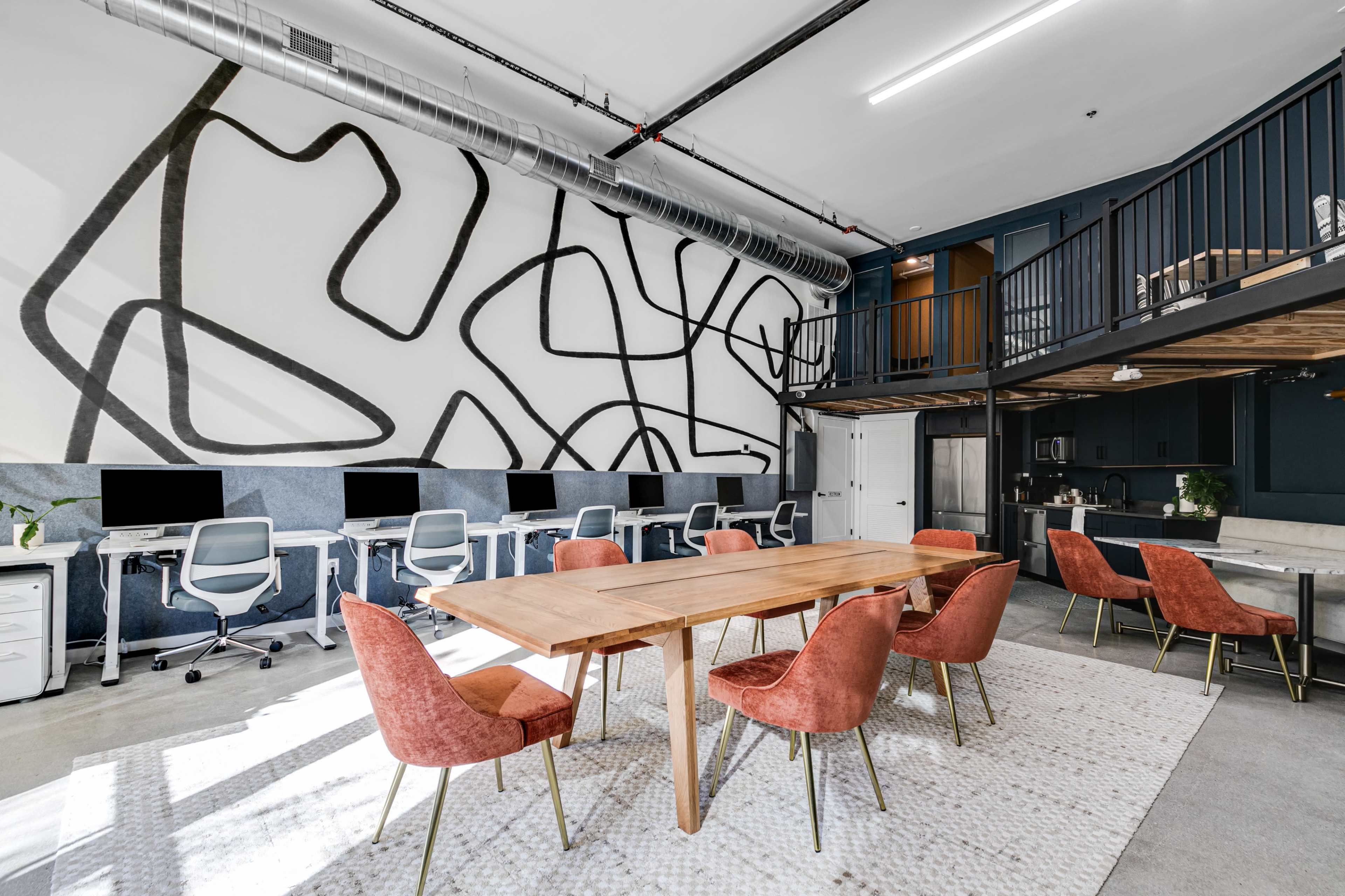 The image shows a modern coworking space with a large wooden table surrounded by pink chairs and workstations featuring computers along one wall.