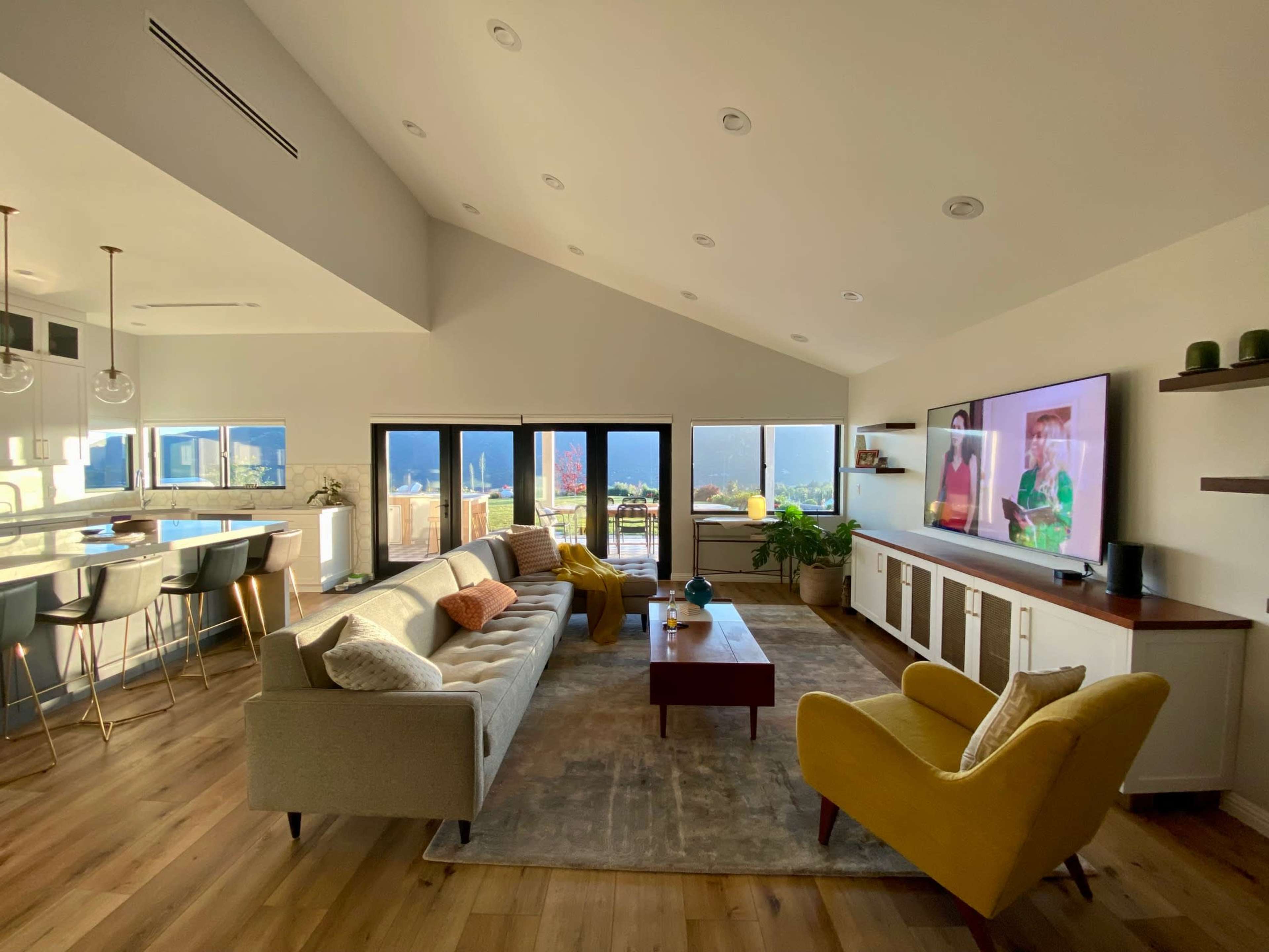 A modern living room features a sectional sofa, an accent chair, a large television, and expansive windows that provide a view of the outdoors.