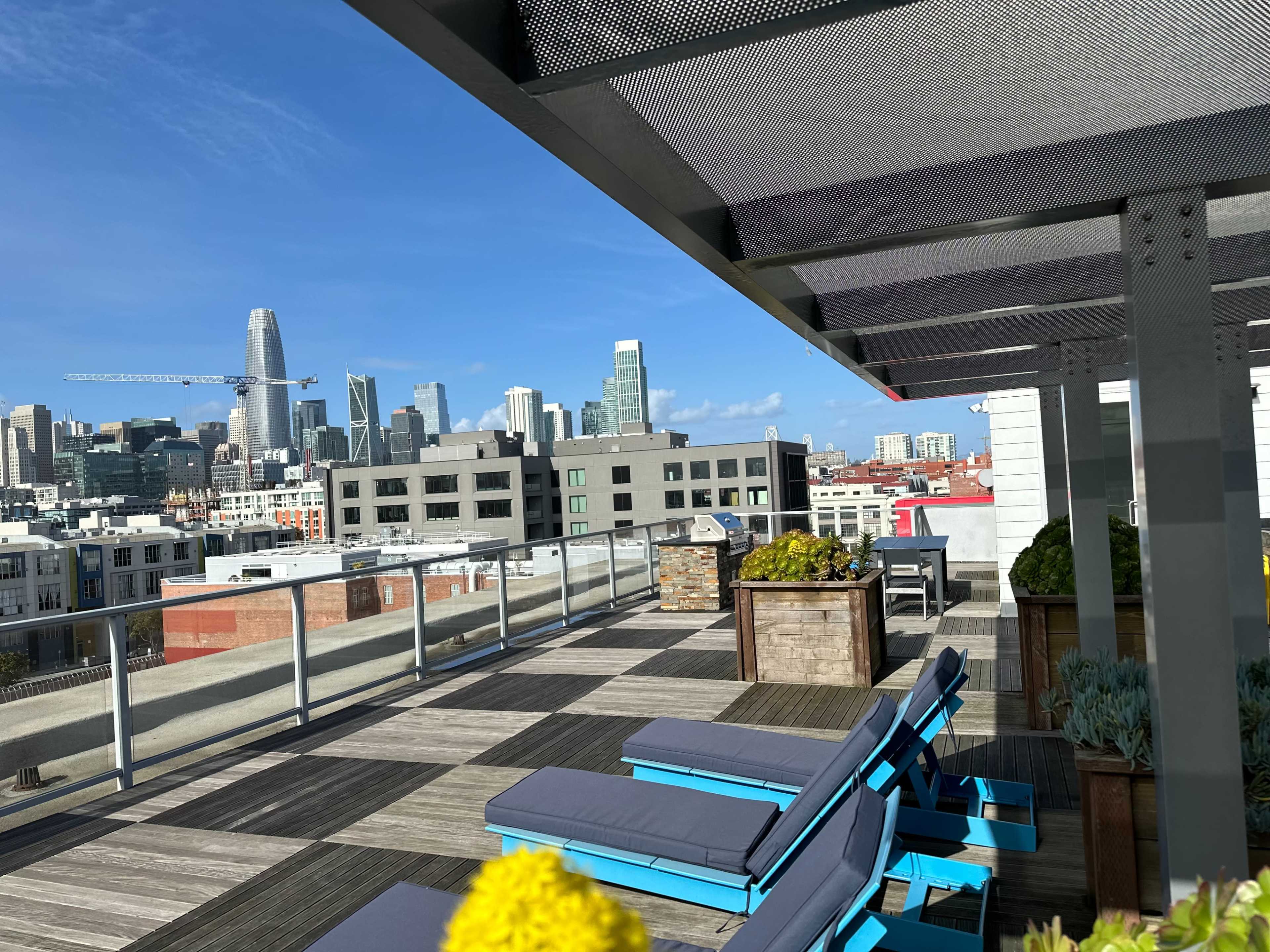 A spacious rooftop terrace with blue lounge chairs, planters, and a view of a city skyline featuring tall buildings and a clear sky.