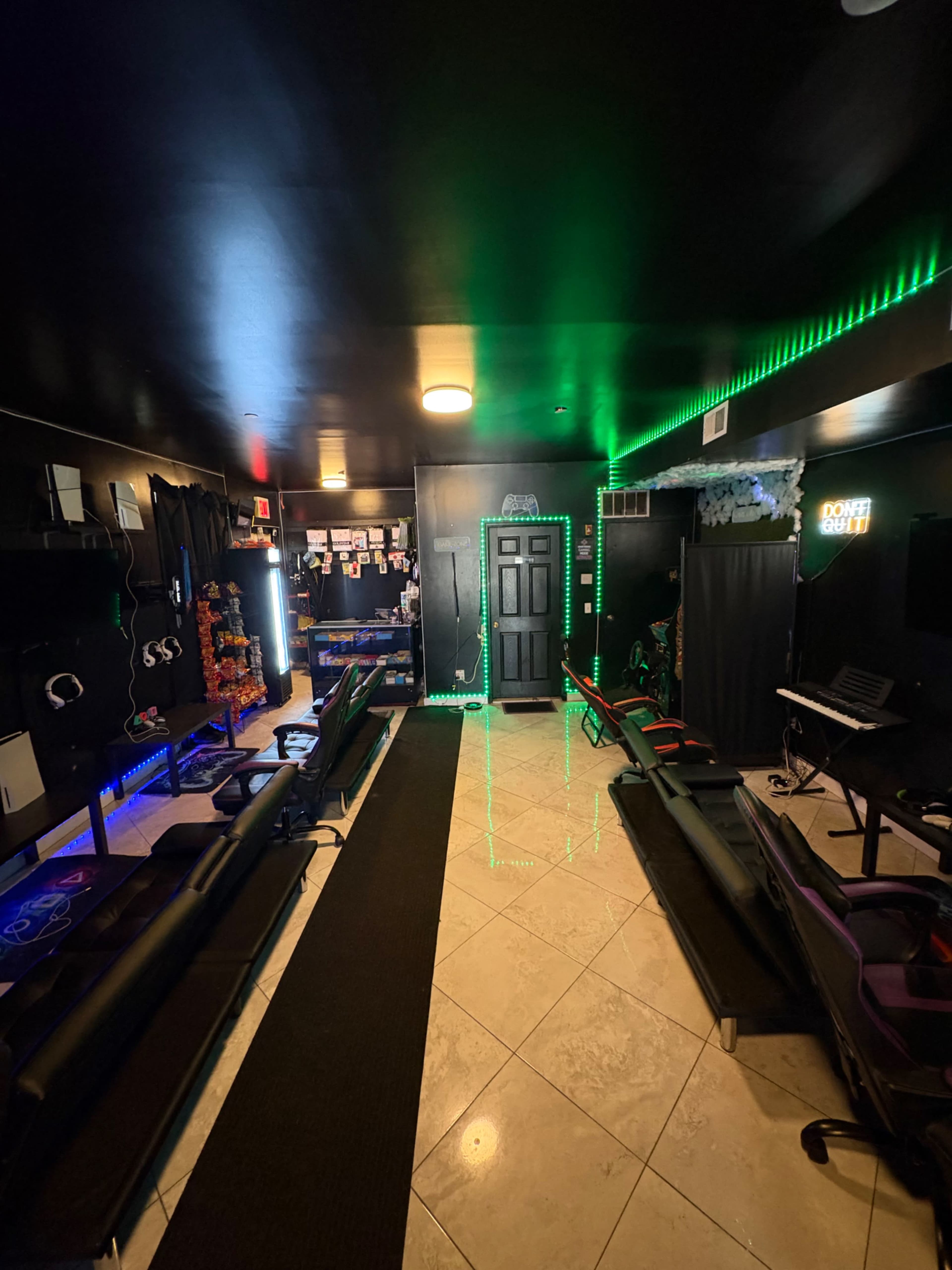 The image shows a gaming lounge with several black leather seating arrangements, neon lighting, and a dark door at the back.