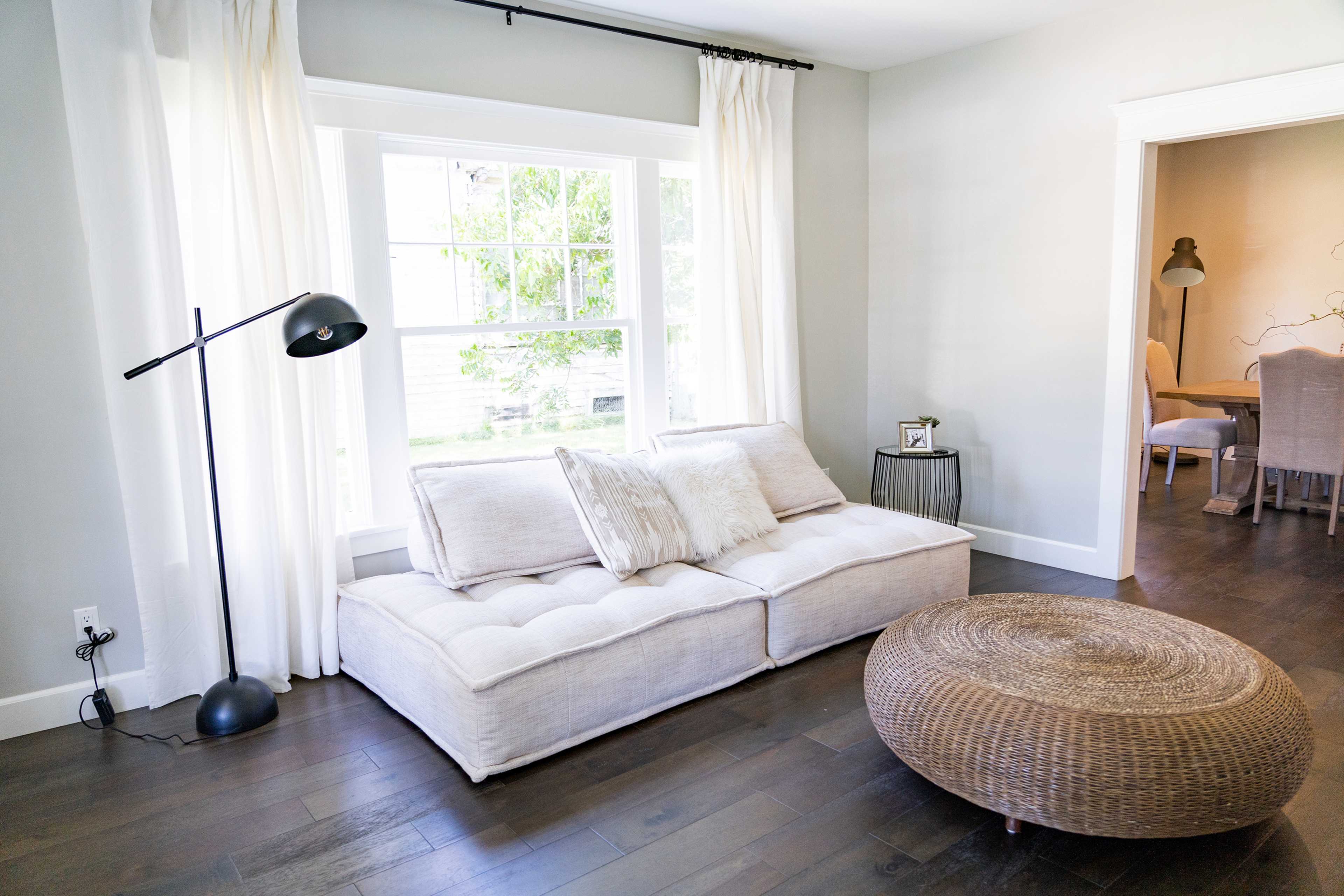 A light-colored sofa with a decorative pillow sits beside a large window, complemented by a round woven coffee table and a standing lamp in a modern living room.