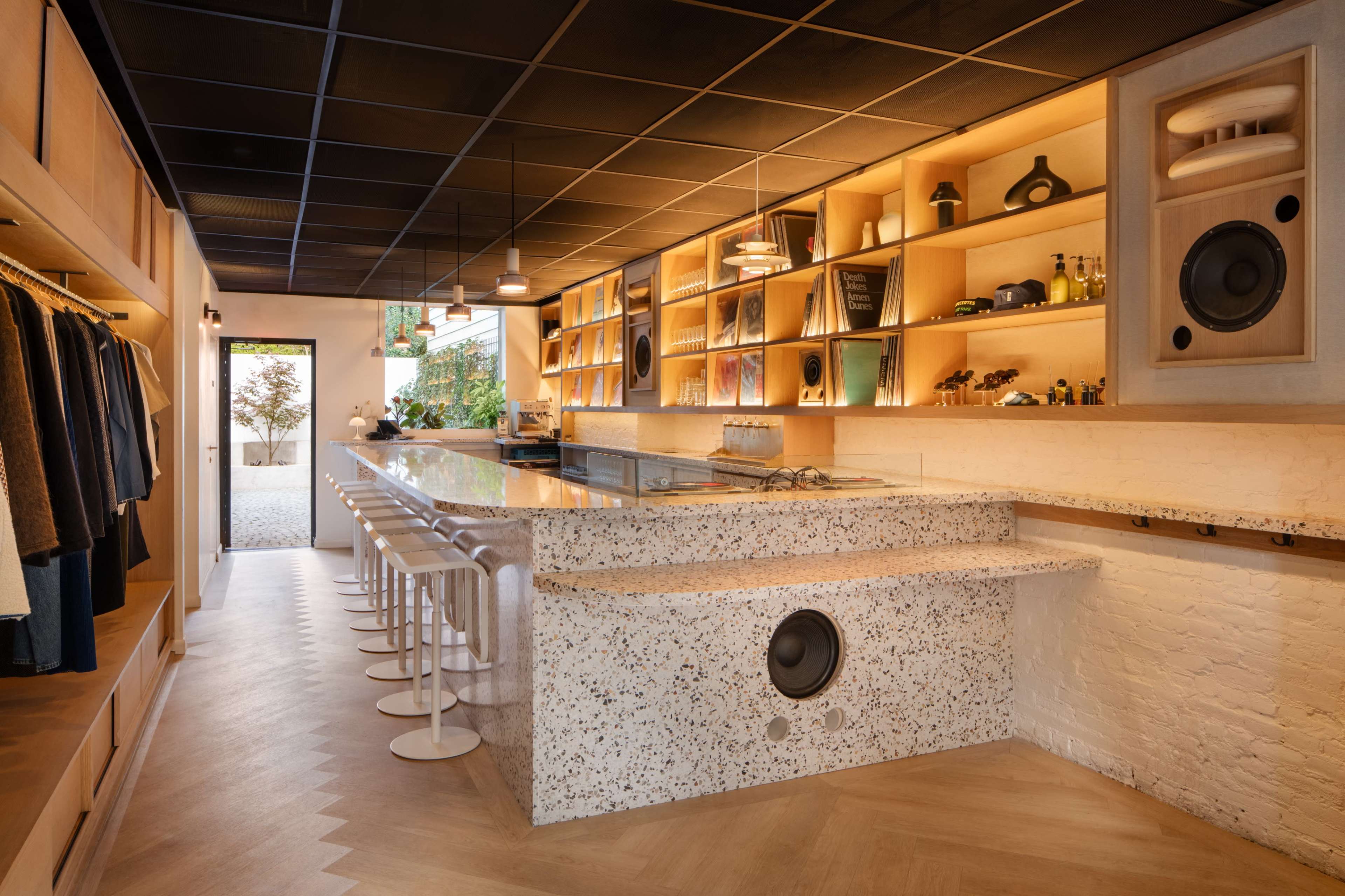 The image shows a modern bar area with a speckled countertop and high stools, alongside wooden shelves displaying various items and an open space in the background featuring greenery.