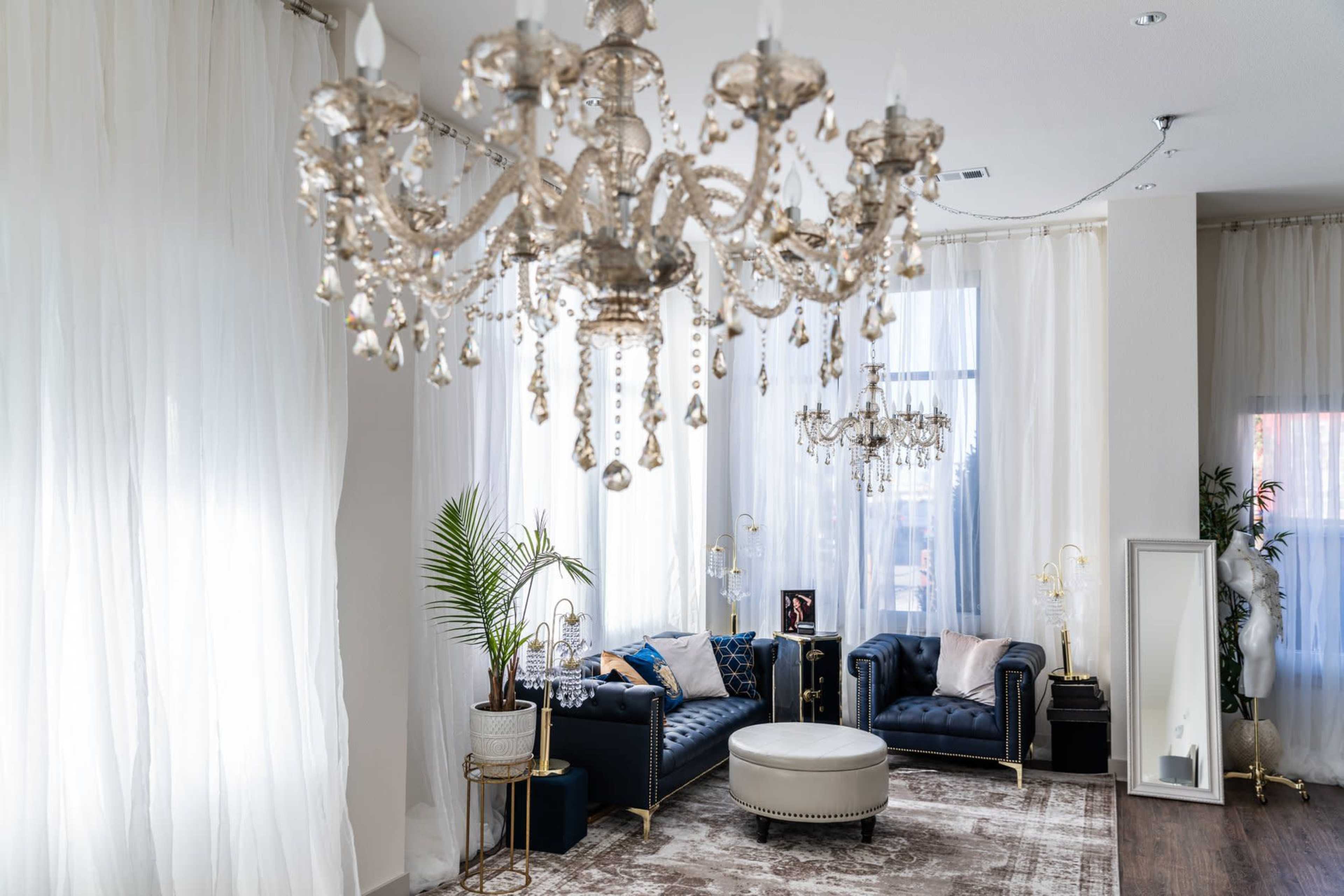 A spacious room features elegant chandeliers above a set of navy blue sofas, a round coffee table, and large windows covered with sheer white curtains.