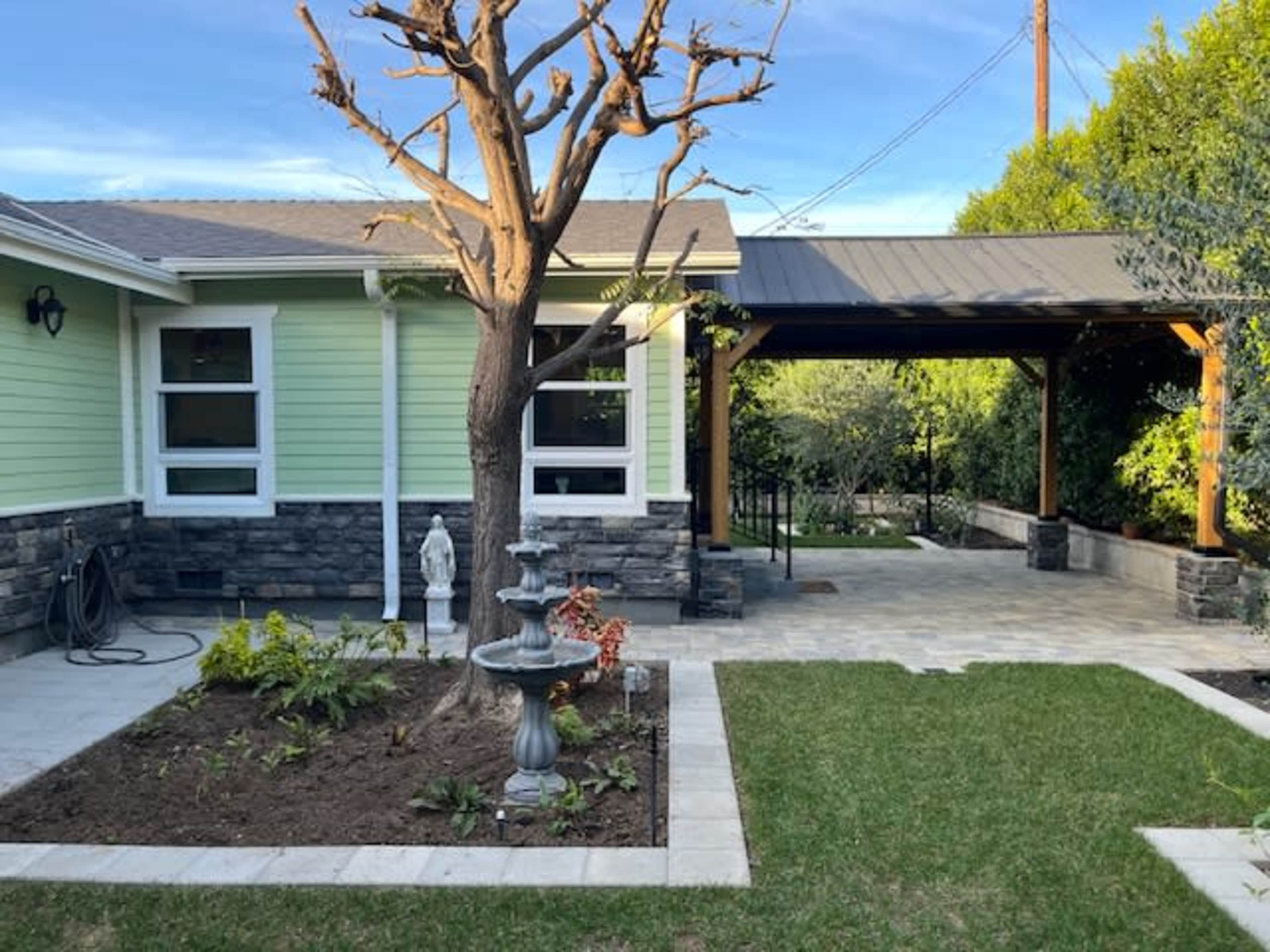 The image shows a landscaped yard with a stone fountain and a dead tree next to a light green house and a covered patio area.