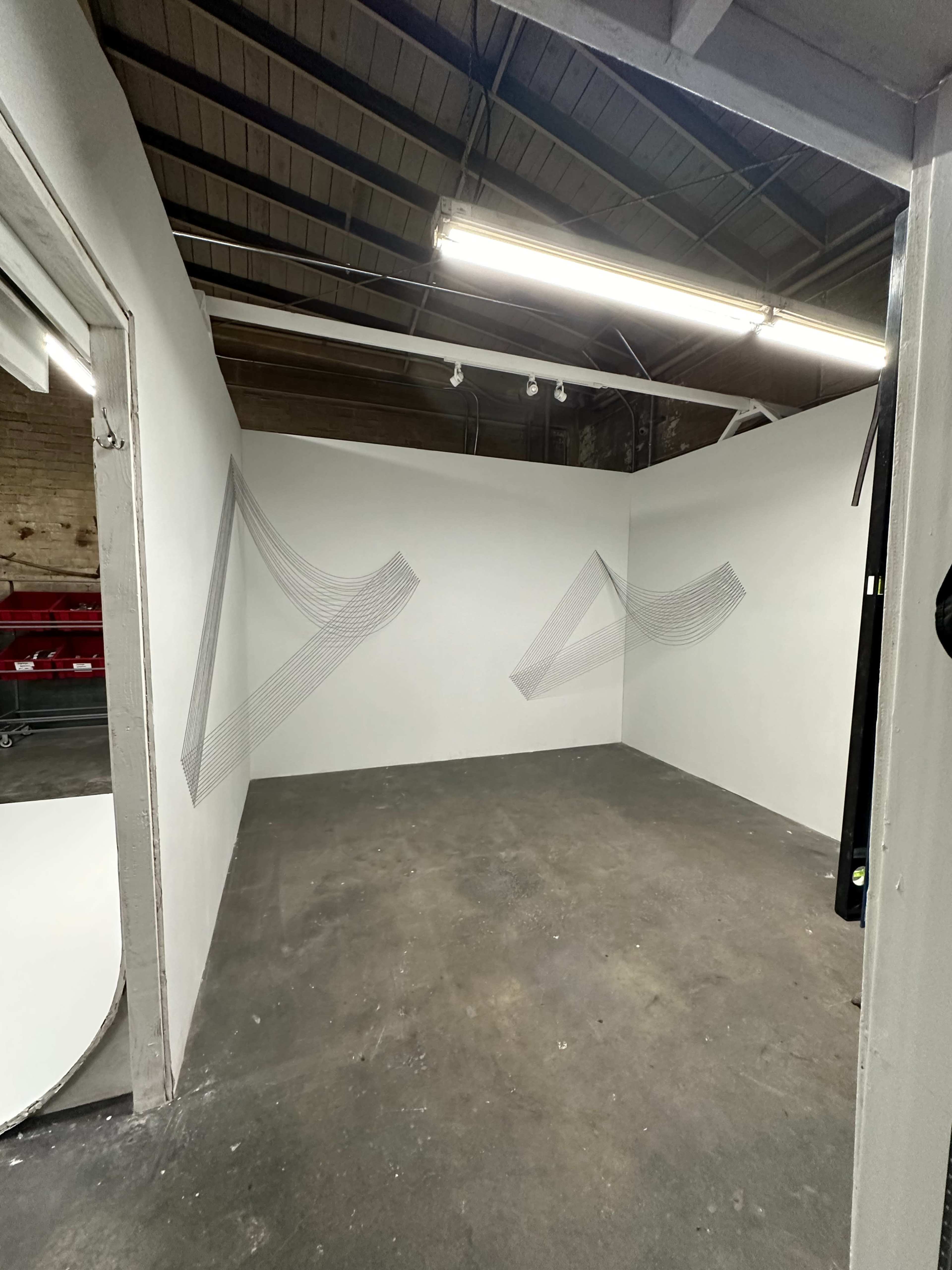 The image shows an empty room with white walls and two geometric wire sculptures mounted on the wall.