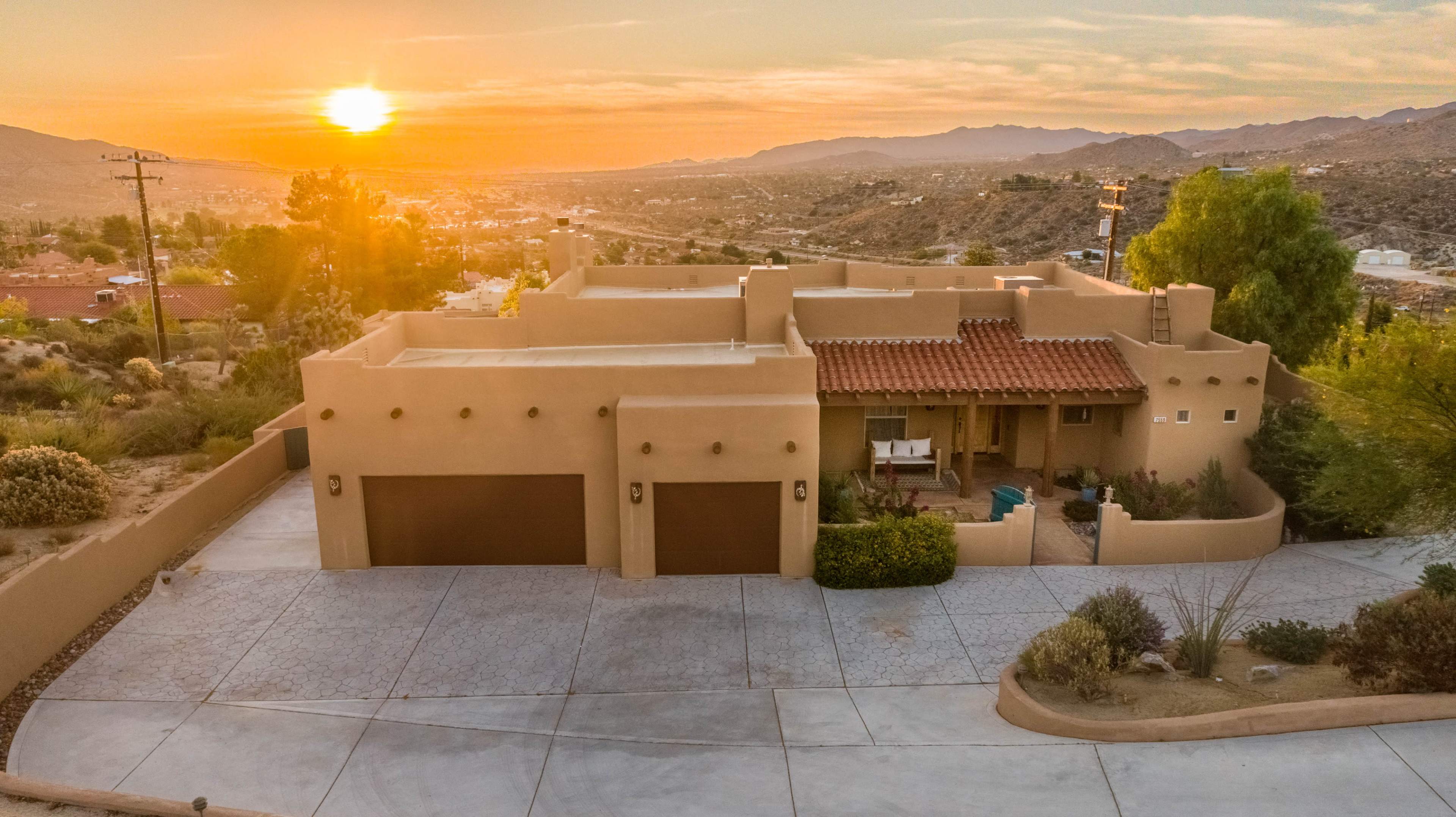 A single-story adobe-style house with two garage doors is set against a sunset view of rolling hills and desert terrain.