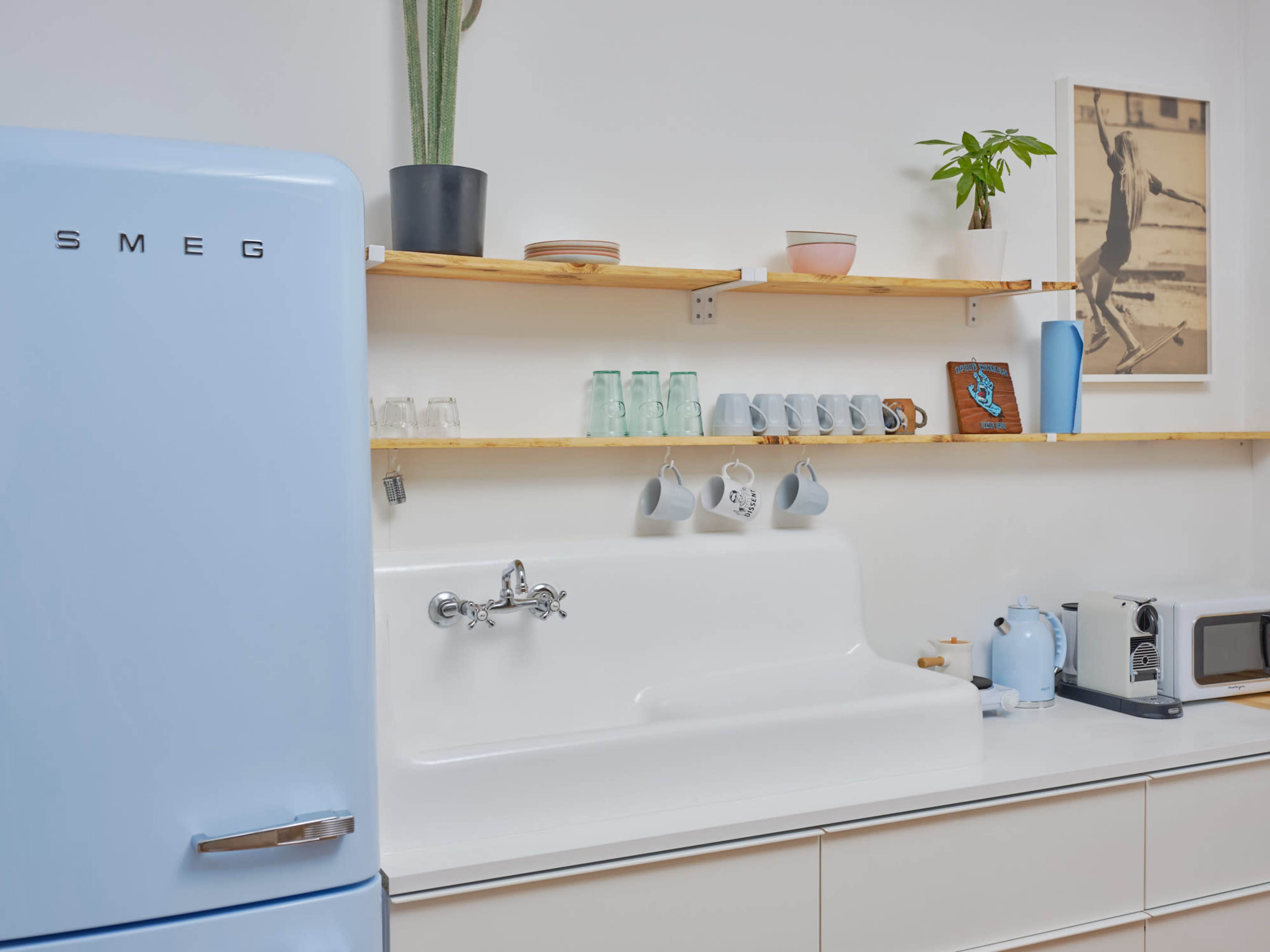 A modern kitchen featuring a light blue SMEG refrigerator, a white porcelain sink, open wooden shelves with various dishes and glasses, and a small countertop microwave and kettle.