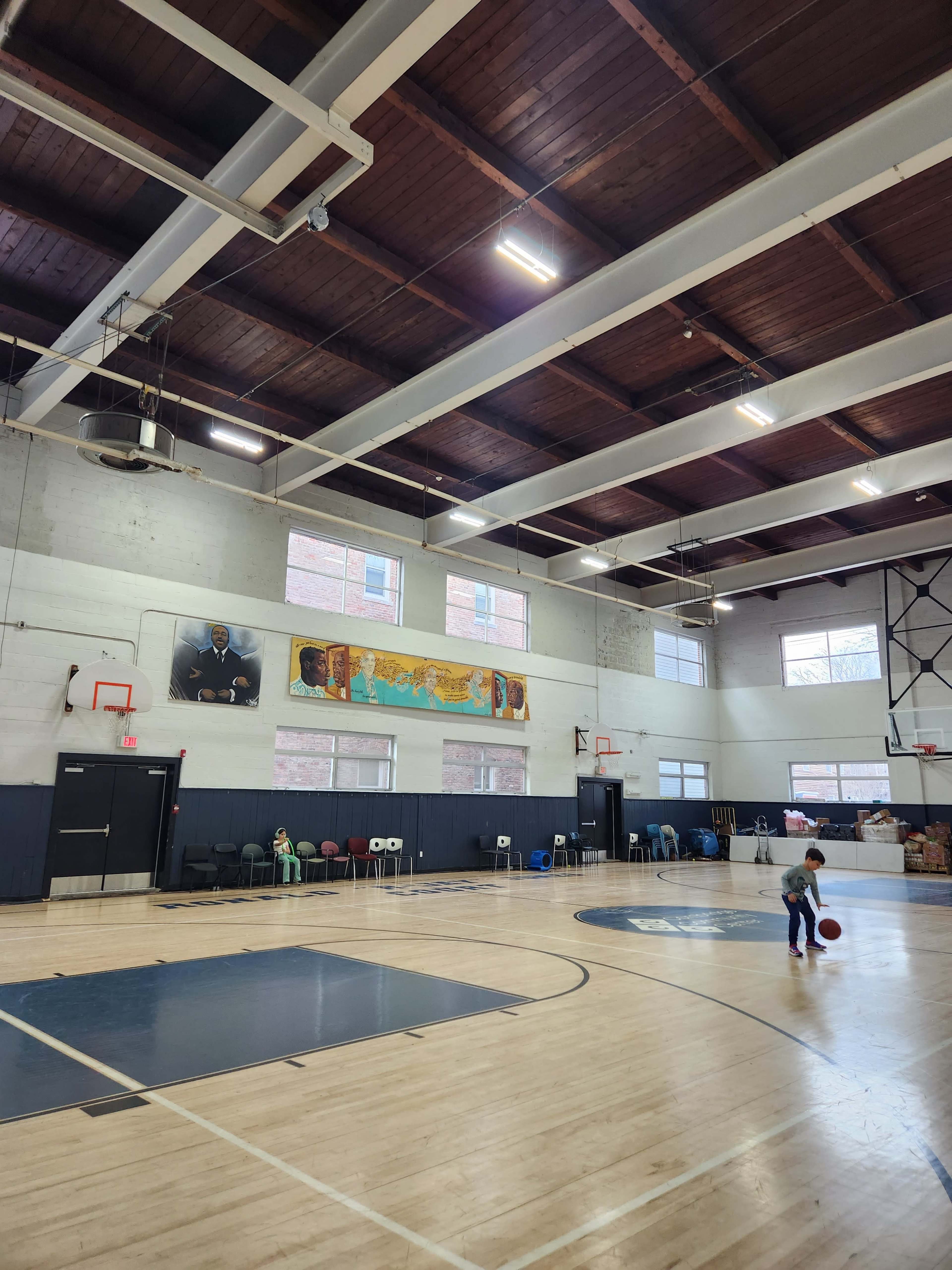 The image shows a gymnasium with a basketball court, high ceilings, and a child playing with a basketball.