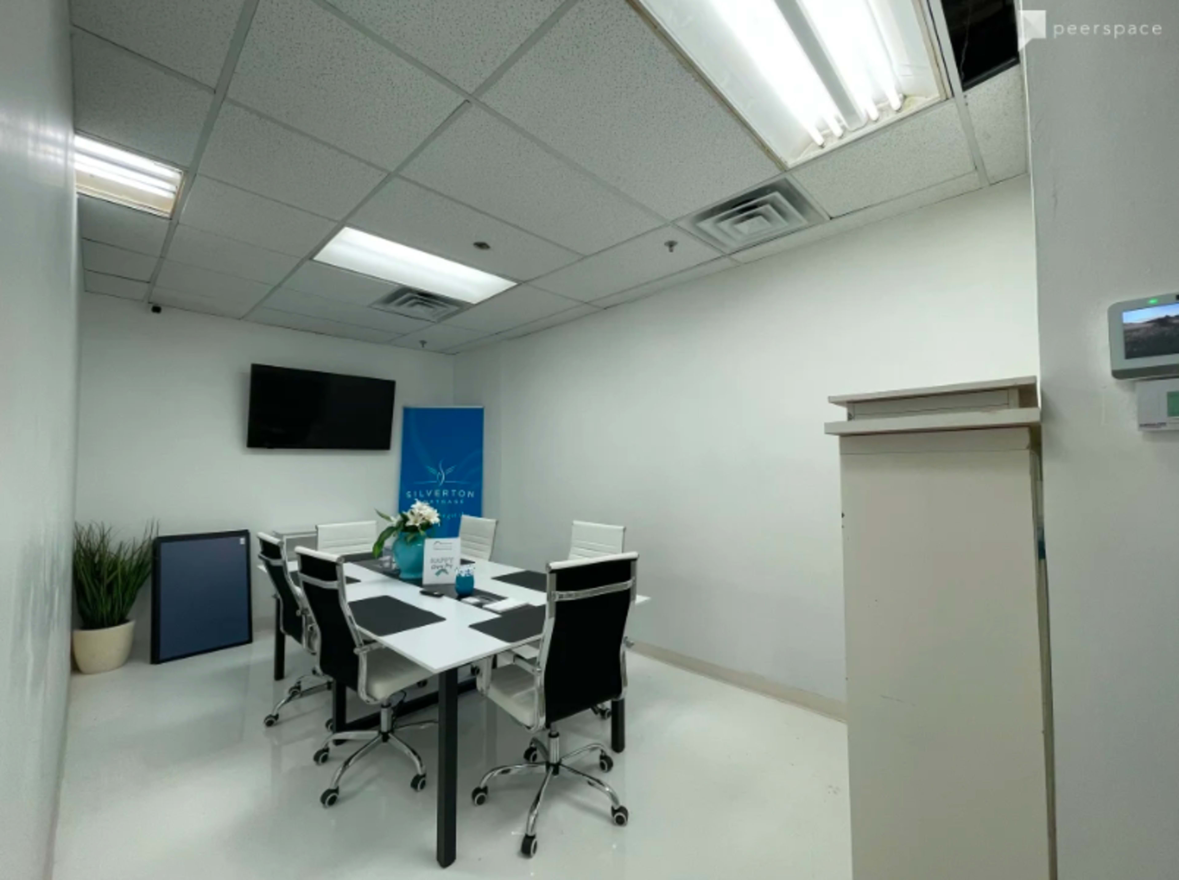 A small, modern conference room with a table and six chairs, a television mounted on the wall, and a plant in a pot.