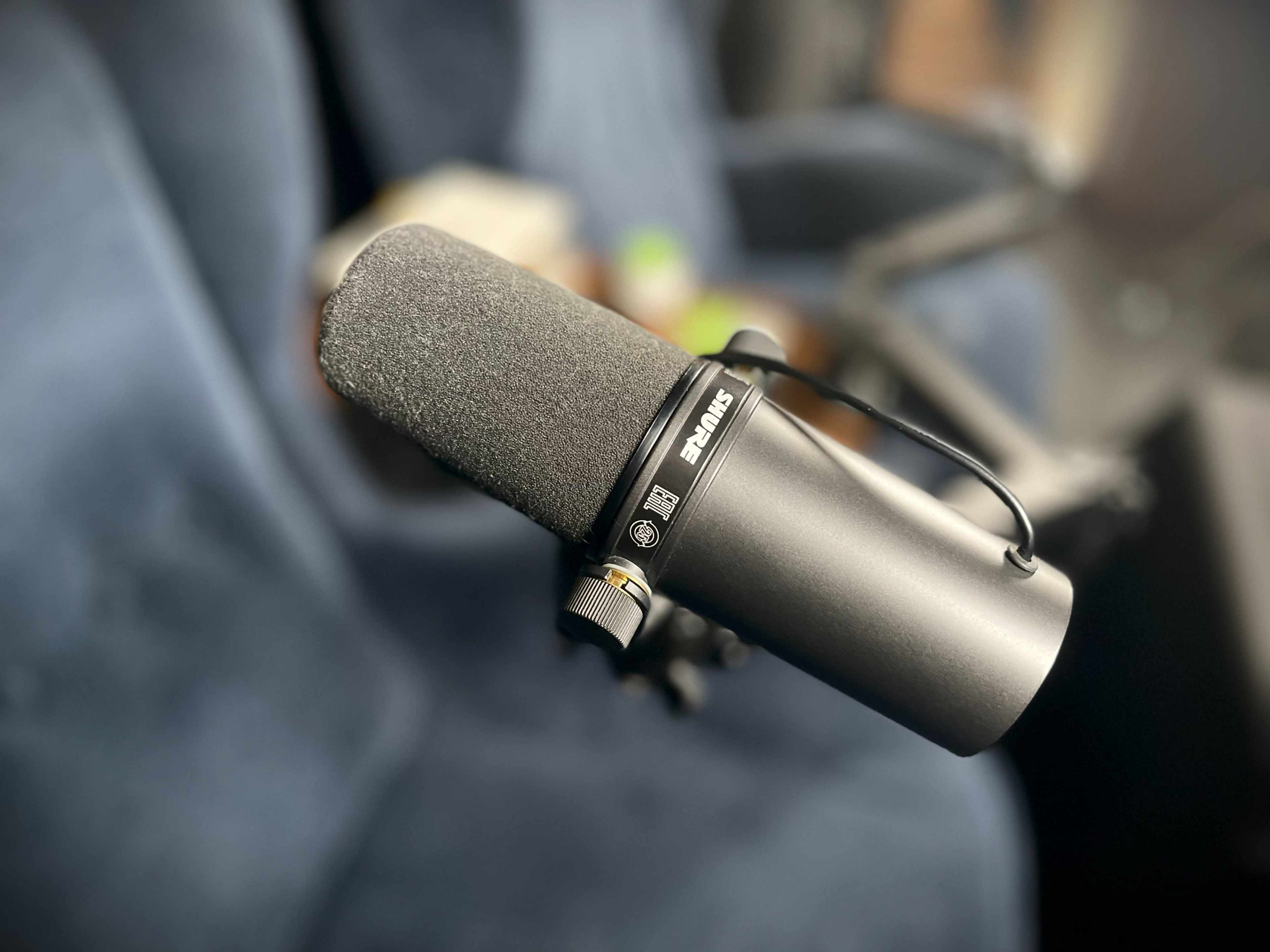 A close-up image of a microphone placed on a stand, set against a blurred background of a seating area.