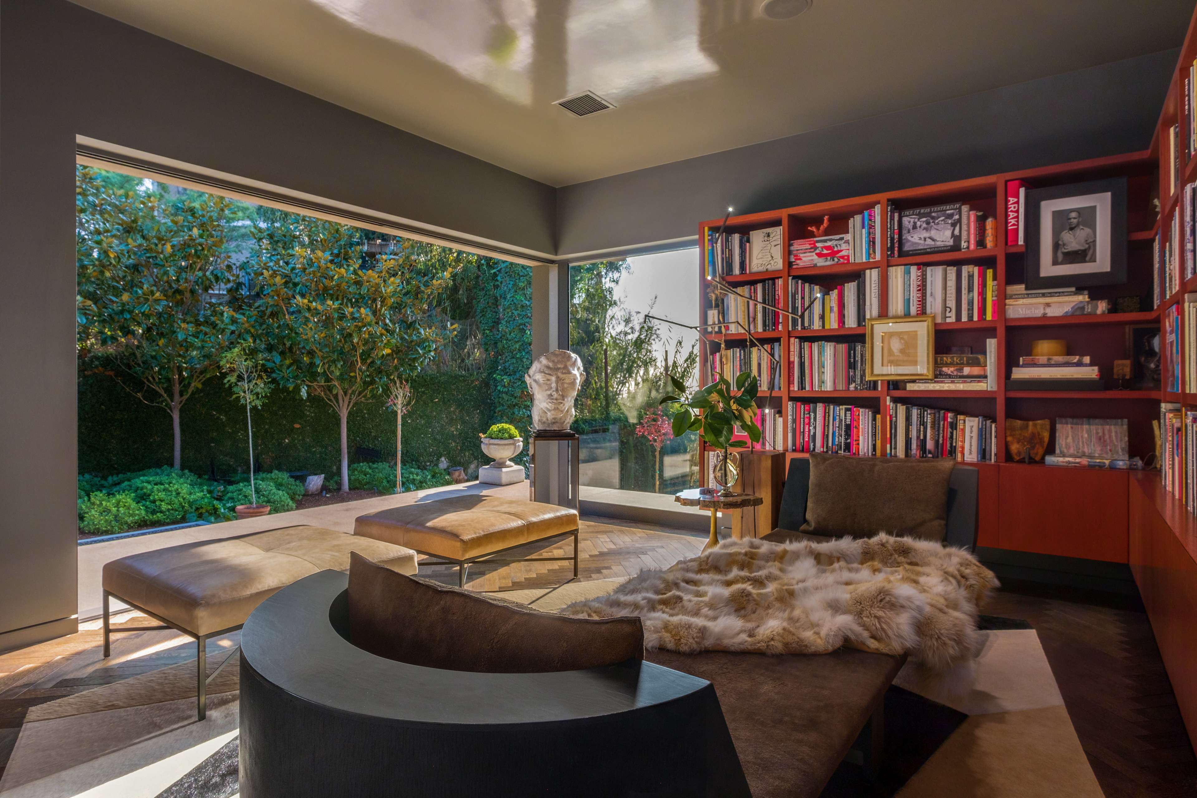 A modern living room features a curved sofa, a bookshelf filled with books, and large windows overlooking a landscaped garden.