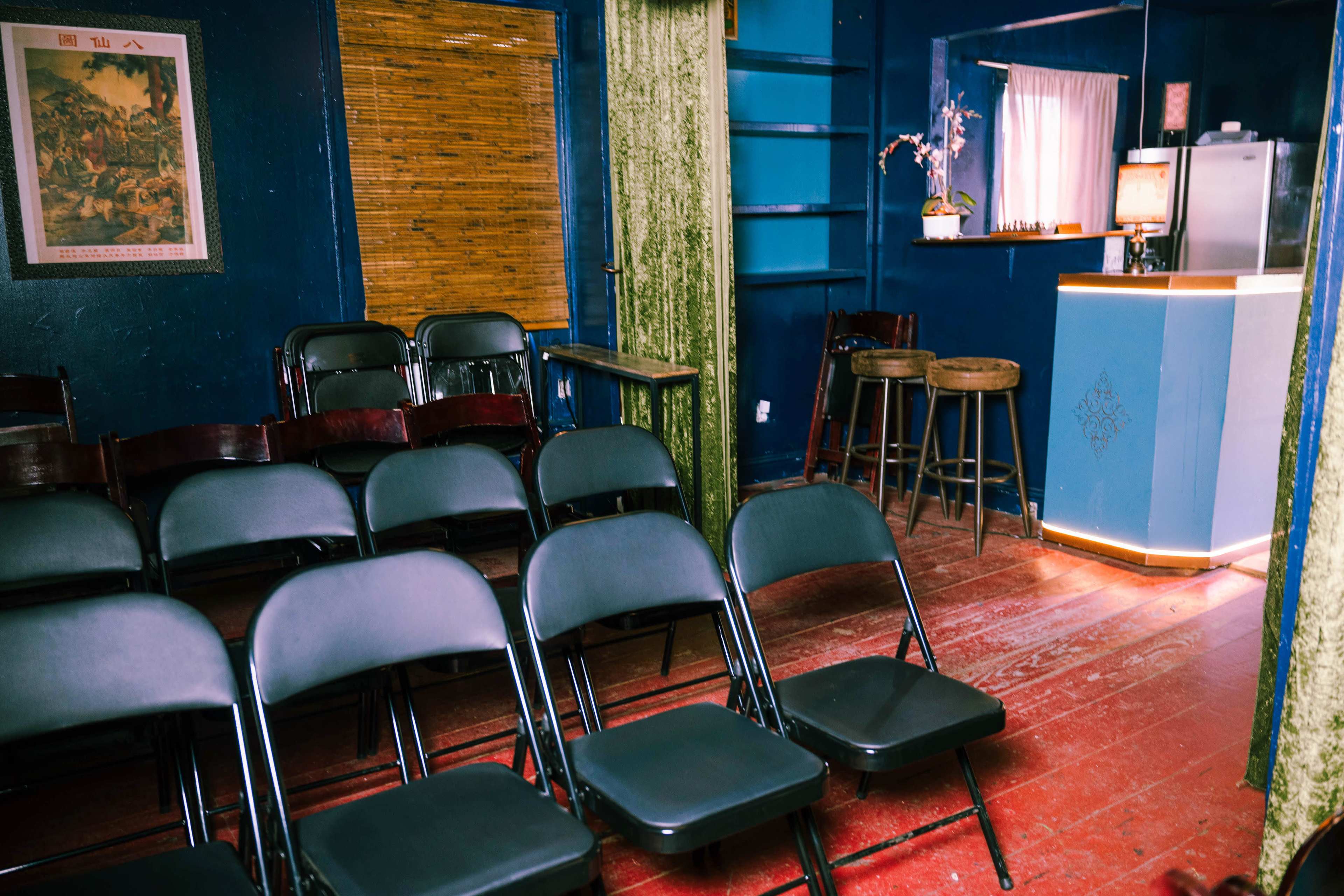 A small, empty room with rows of black folding chairs facing a bar area illuminated by soft lighting.