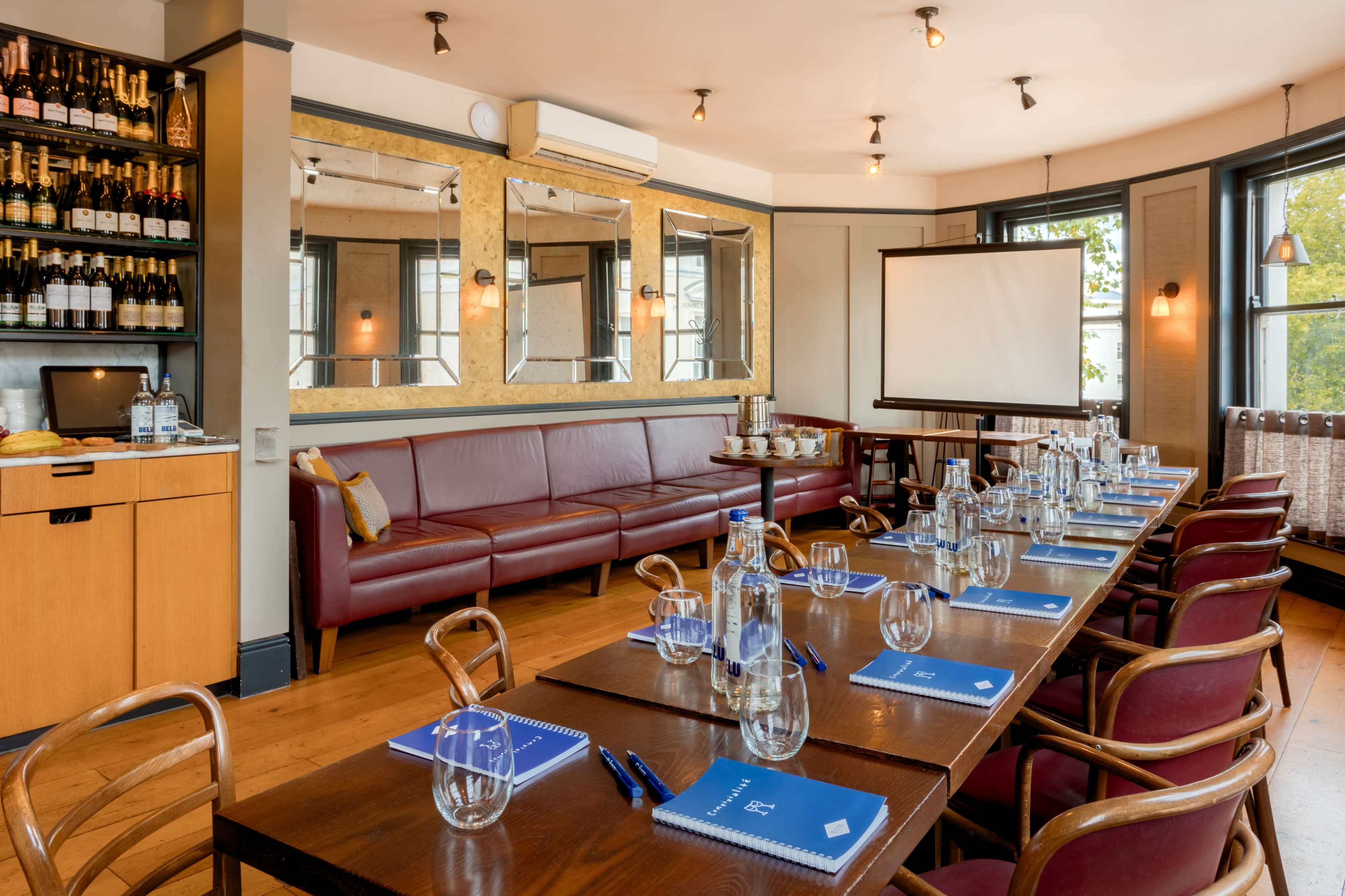 A conference room features a long wooden table set with glasses and notebooks, surrounded by chairs, and includes a large screen and a bar area stocked with beverages.