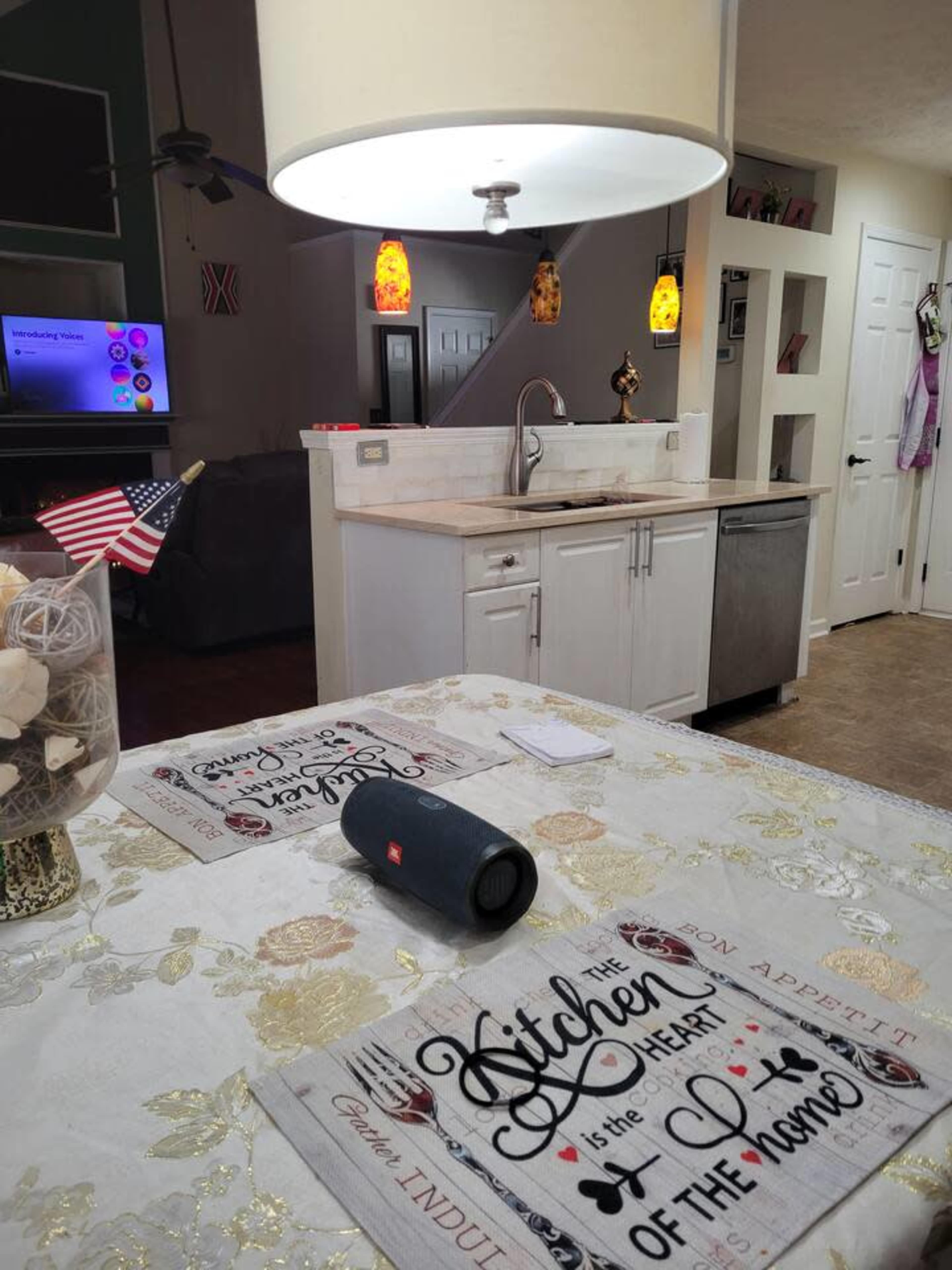 A kitchen scene features a table with decorative placemats, a Bluetooth speaker, and a flag, with a sink and appliances visible in the background.