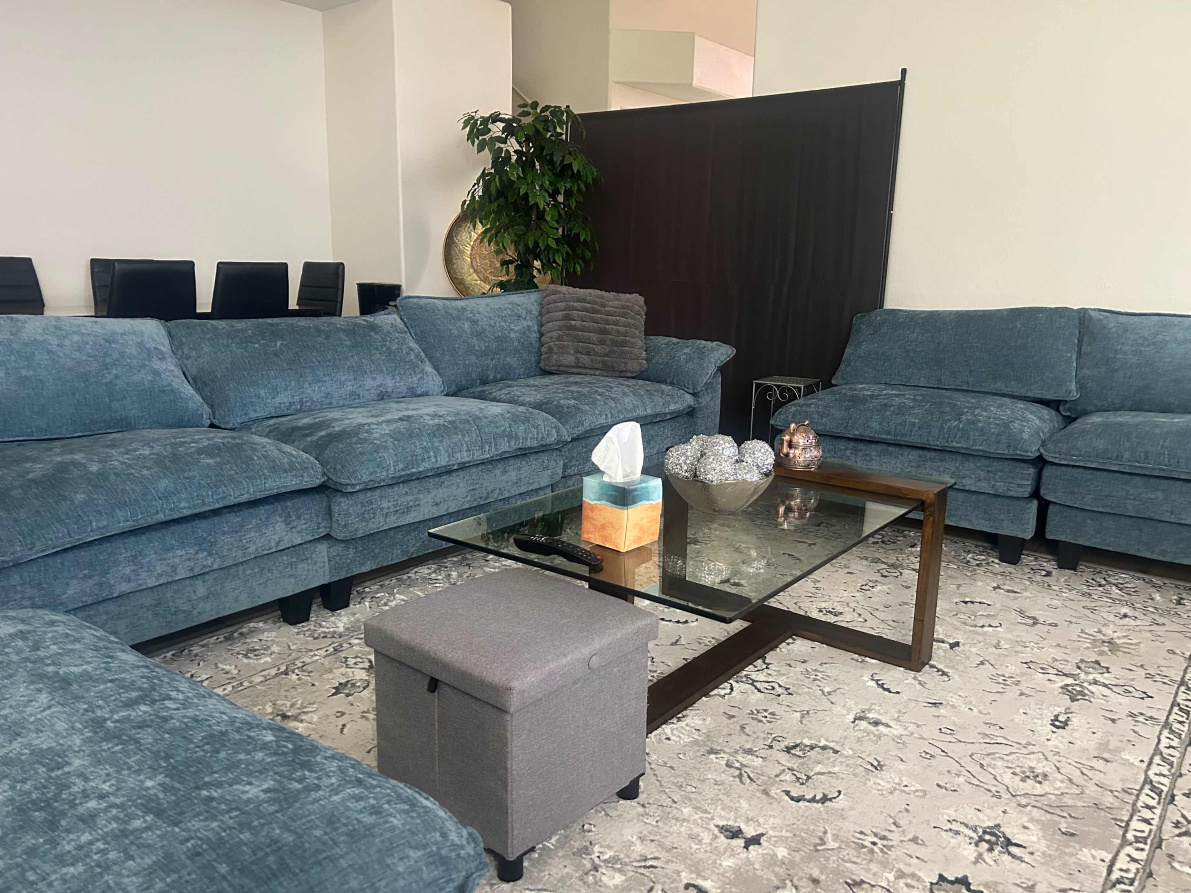 The image shows a living room with blue plush sofas arranged around a glass coffee table, featuring a decorative pouch and a few ornaments on top, set on a patterned rug.