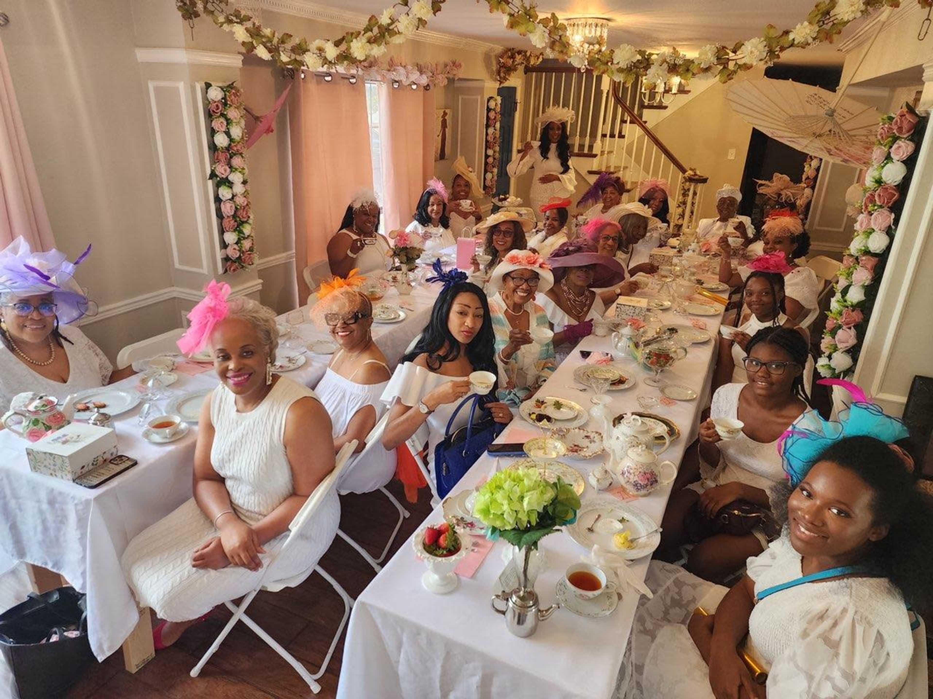 A group of women wearing white dresses and colorful hats gathers around a long table set for a tea party.