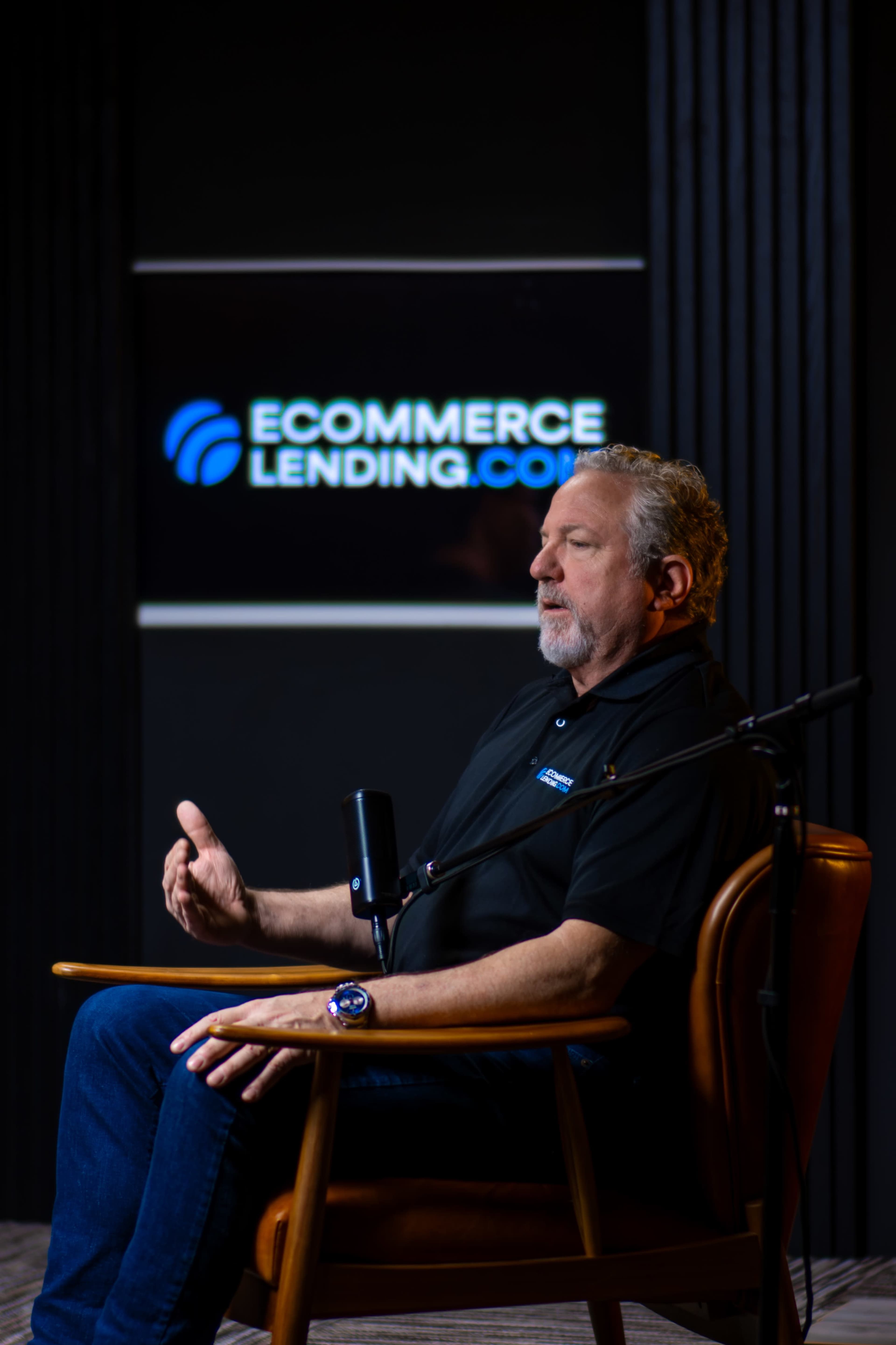 A man sits in a chair, gesturing while speaking, in front of a backdrop featuring the logo of Ecommerce Lending.