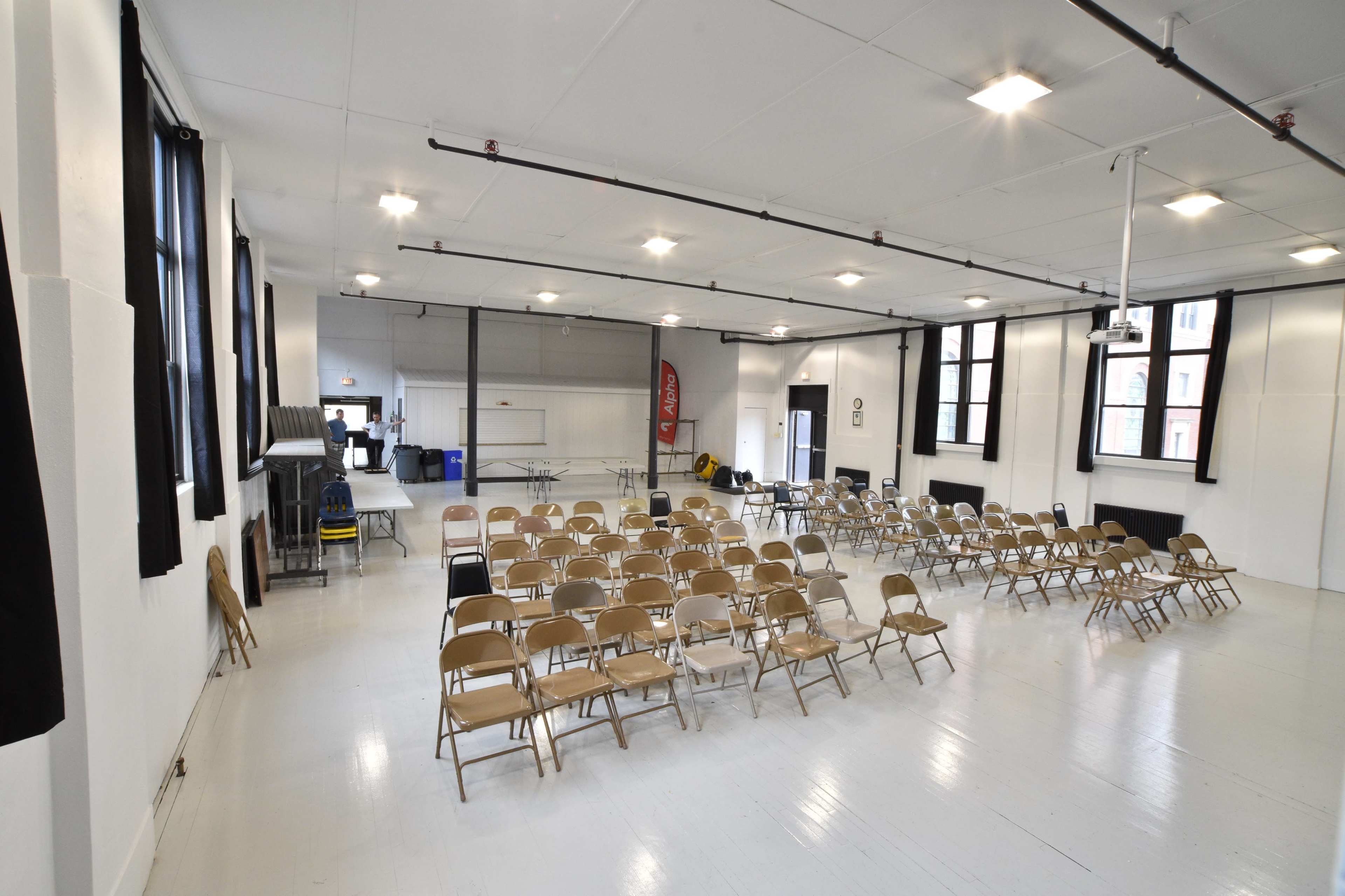 A spacious indoor area set up with rows of folding chairs facing a projection screen and a table along one side.