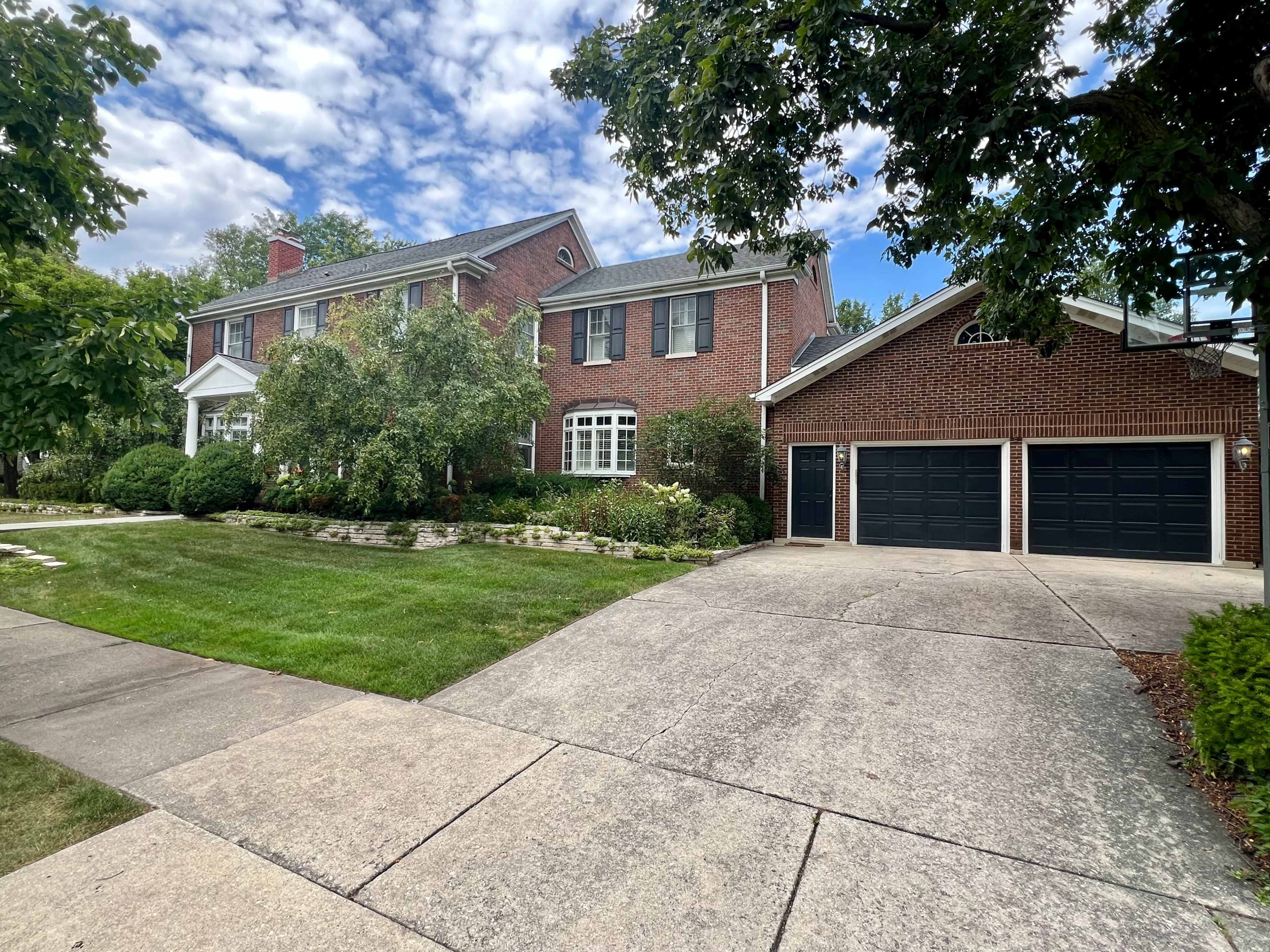 A brick two-story house with a landscaped yard and a two-car garage stands adjacent to a concrete driveway.