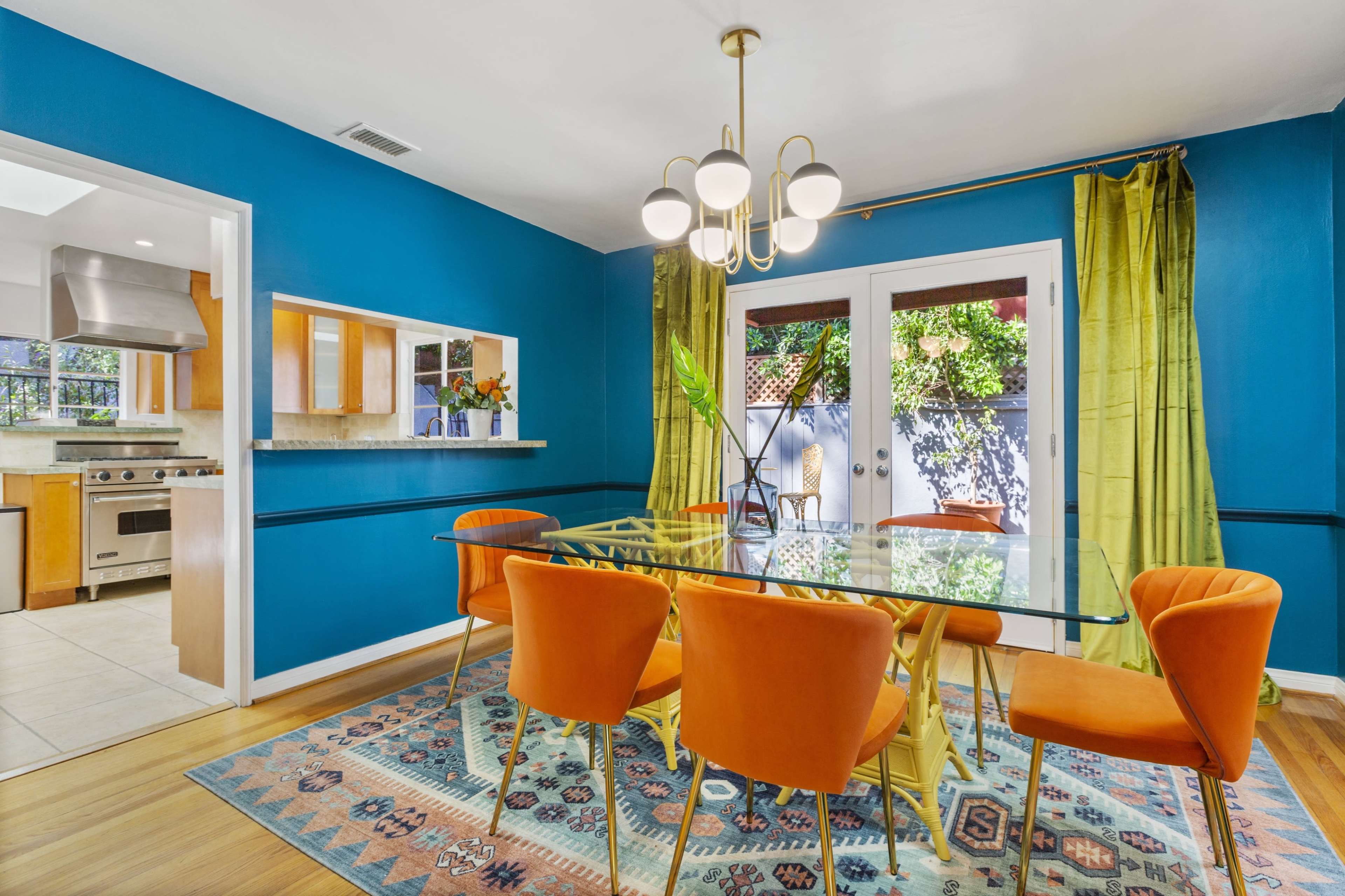 A dining area with a glass table surrounded by orange chairs, accented by blue walls and green curtains.