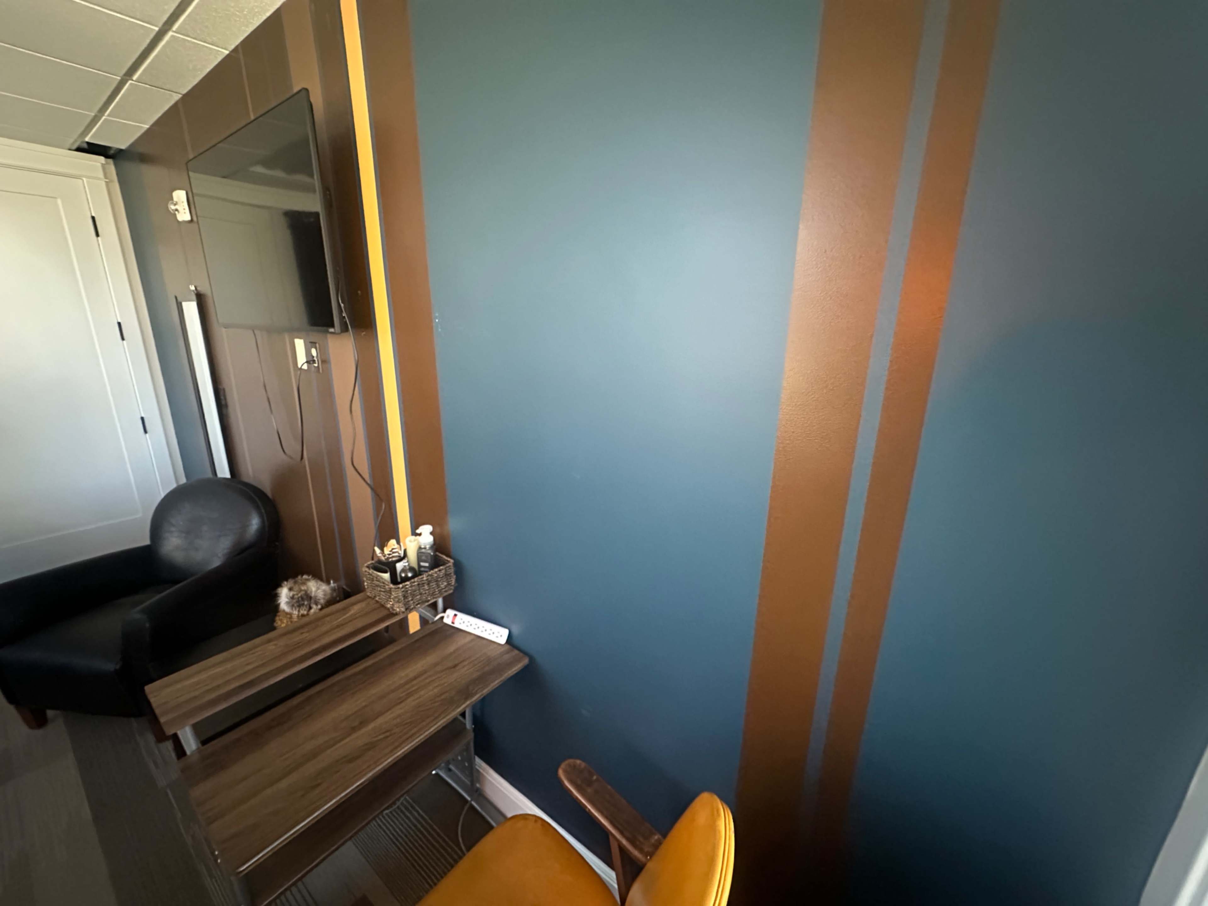 The image shows a small room with a teal wall featuring vertical brown stripes, a wooden desk, a yellow chair, and a black armchair in the corner.