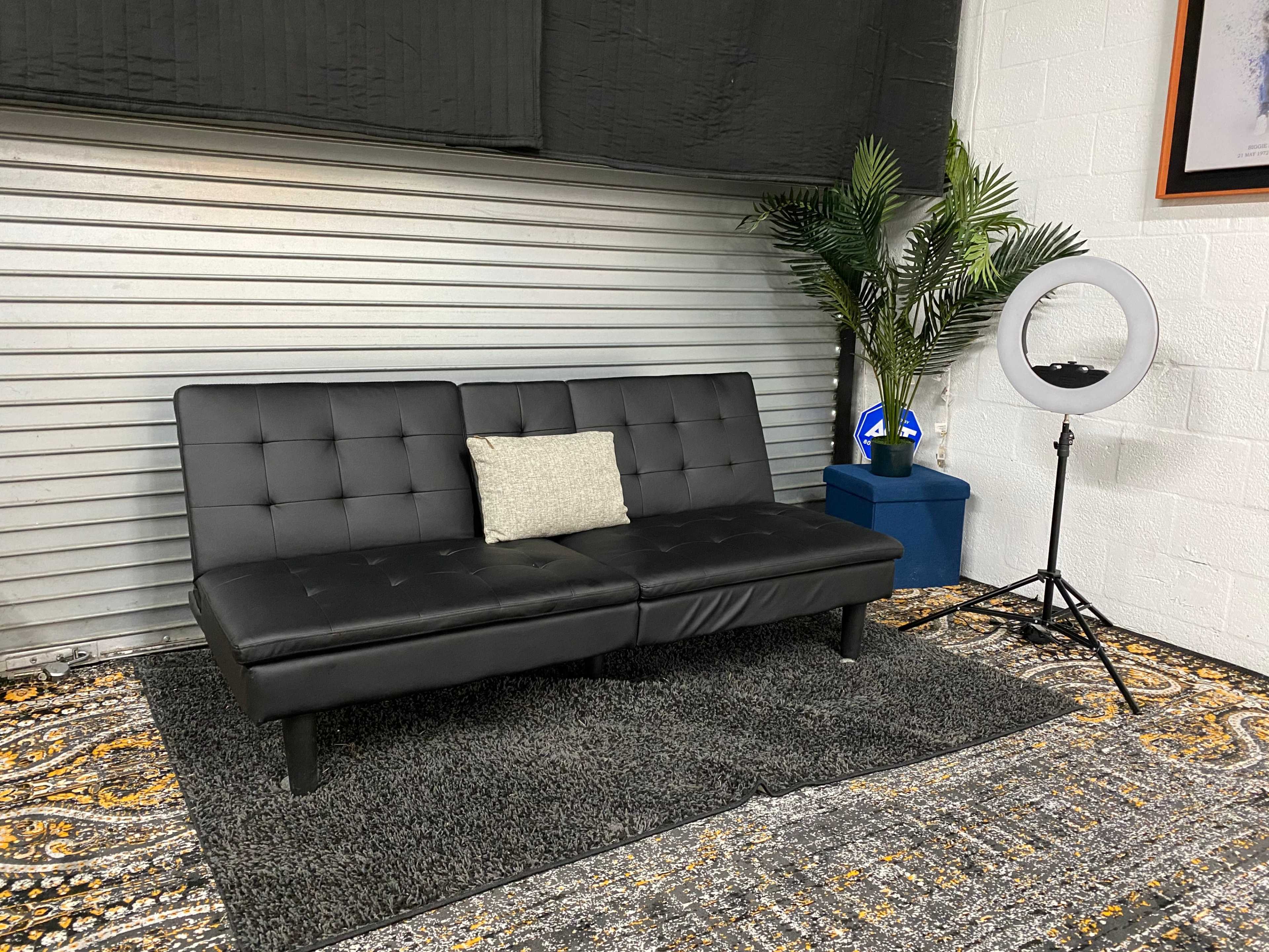 A black futon is positioned on a patterned rug in front of a metal backdrop, accompanied by a floor plant and a ring light on a stand.