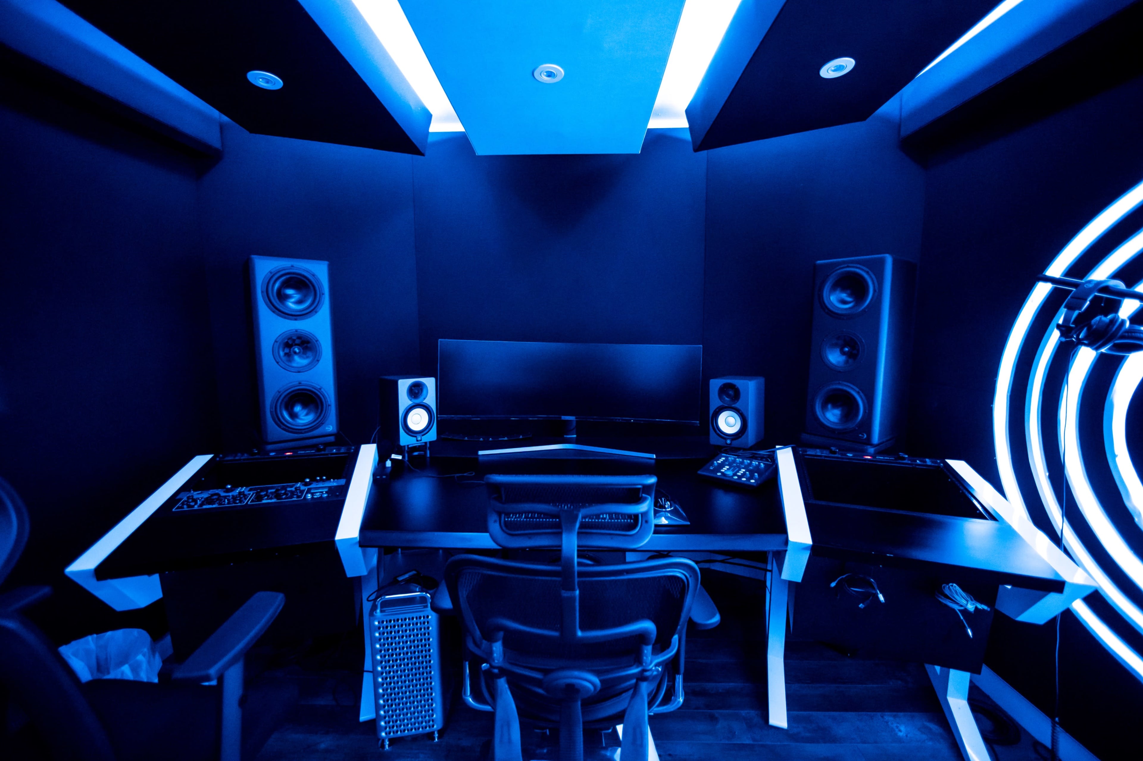 A modern recording studio features a sleek black desk with multiple audio equipment, surrounded by large speakers and illuminated by blue lighting.