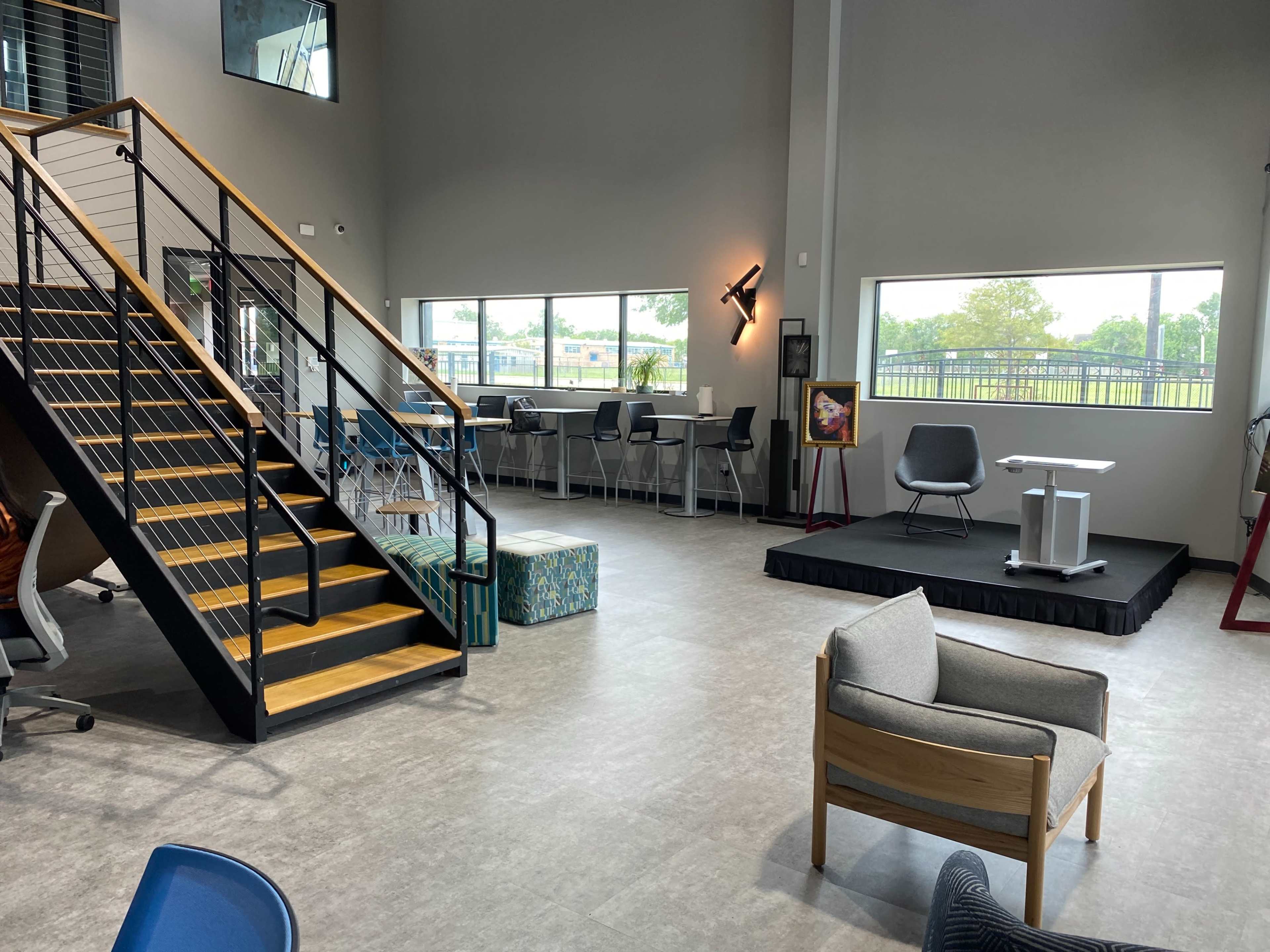 The image shows a modern interior space featuring a staircase, seating areas, workstations, and a stage-like platform.