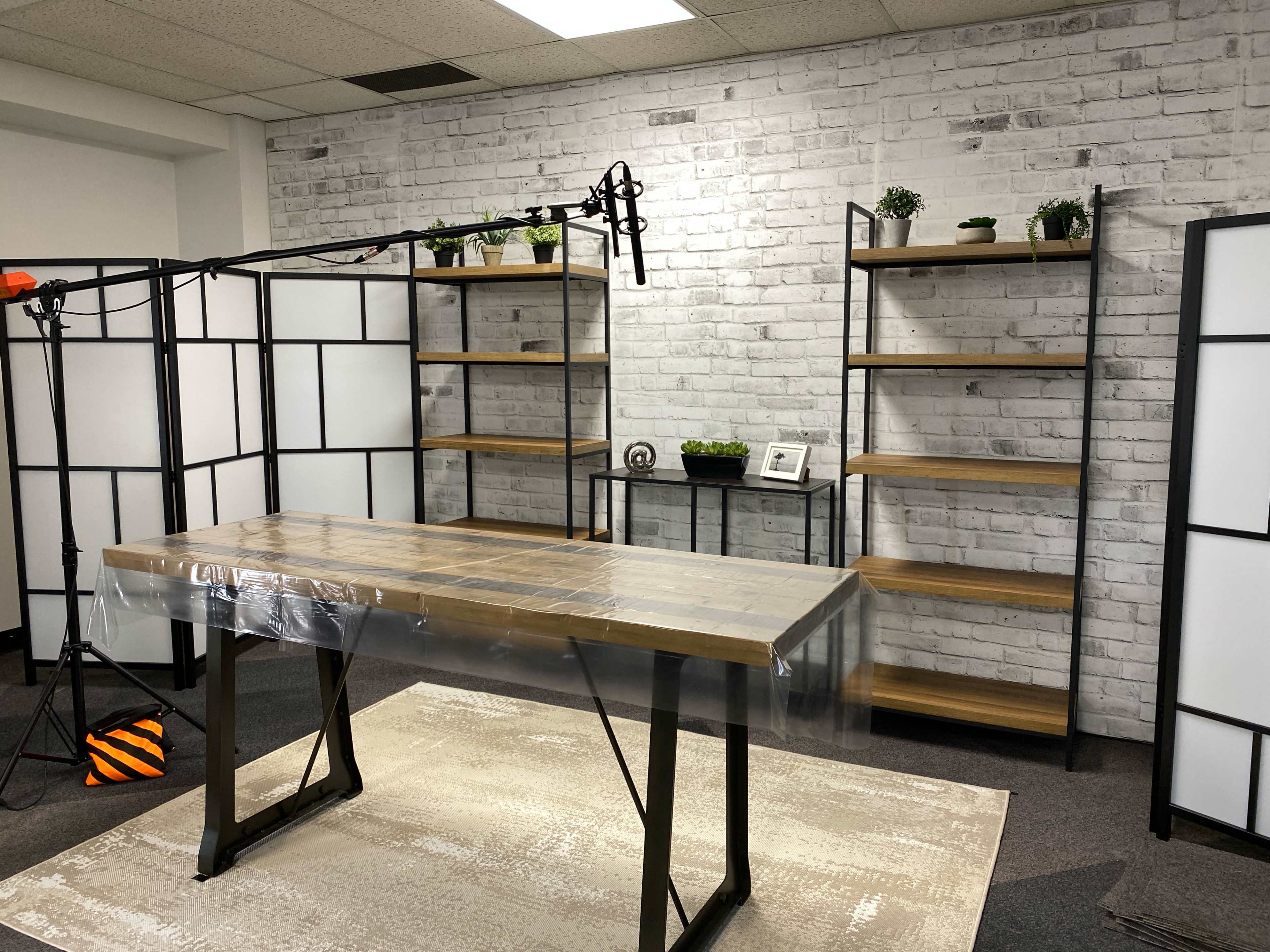 A studio setup features a wooden table covered with plastic in front of black metal shelves filled with small plants and decorative items, against a white brick wall.
