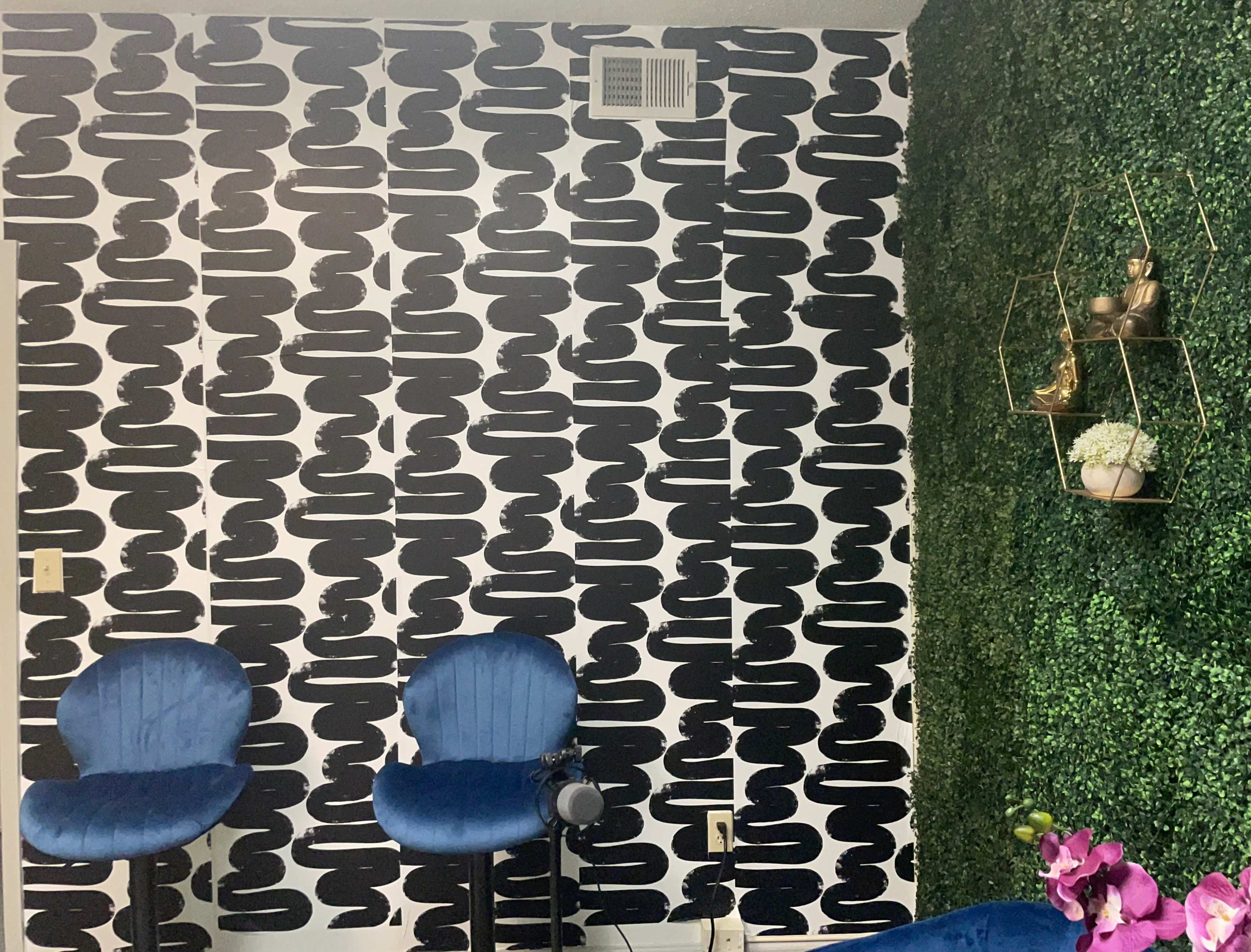A room with a wall covered in a black-and-white patterned wallpaper, two blue chairs in front of it, and a section of artificial green wall with decorative shelves.