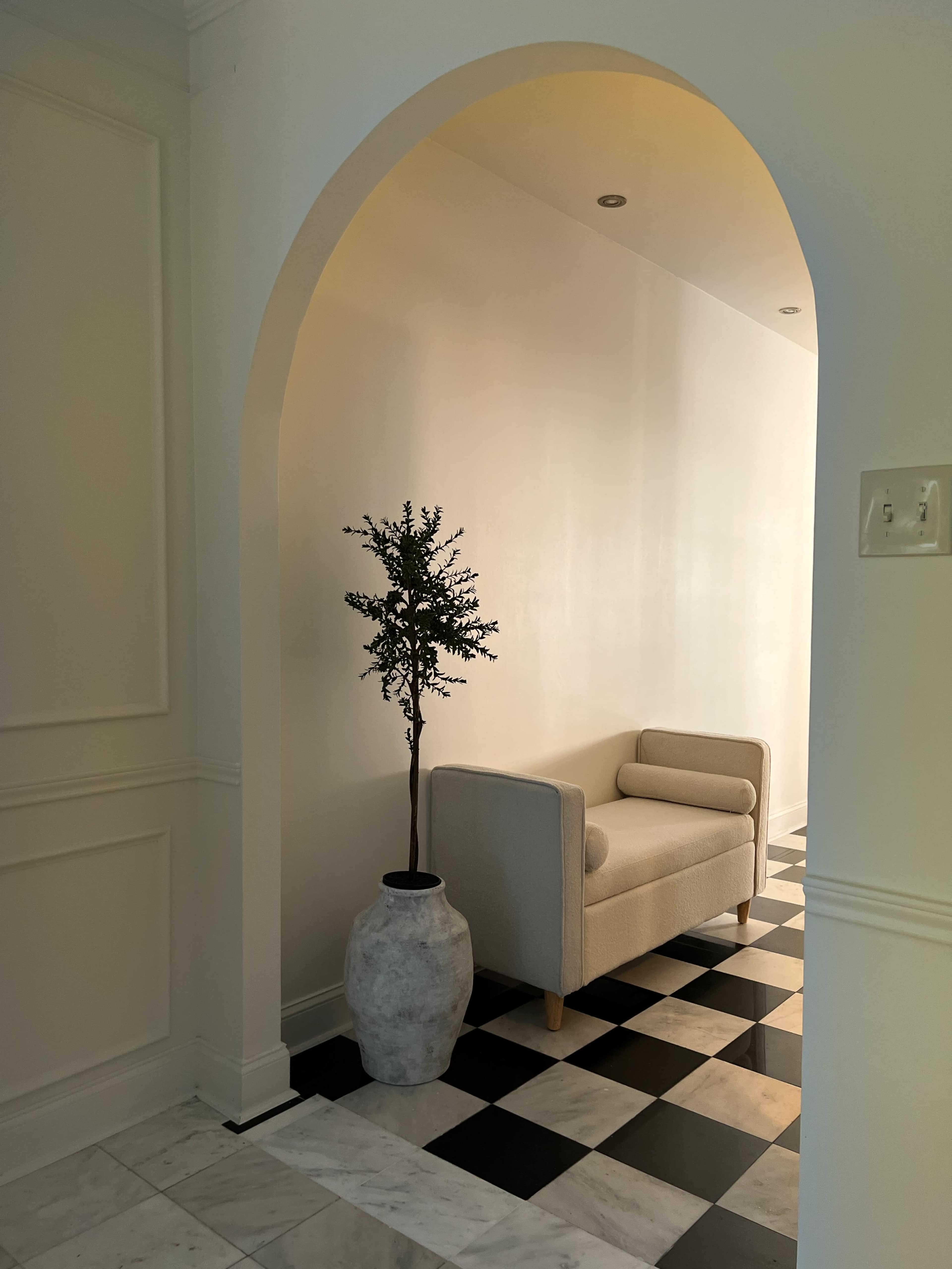 A cream-colored sofa is positioned beside a potted plant within an archway leading to a hallway with black and white tiled flooring.