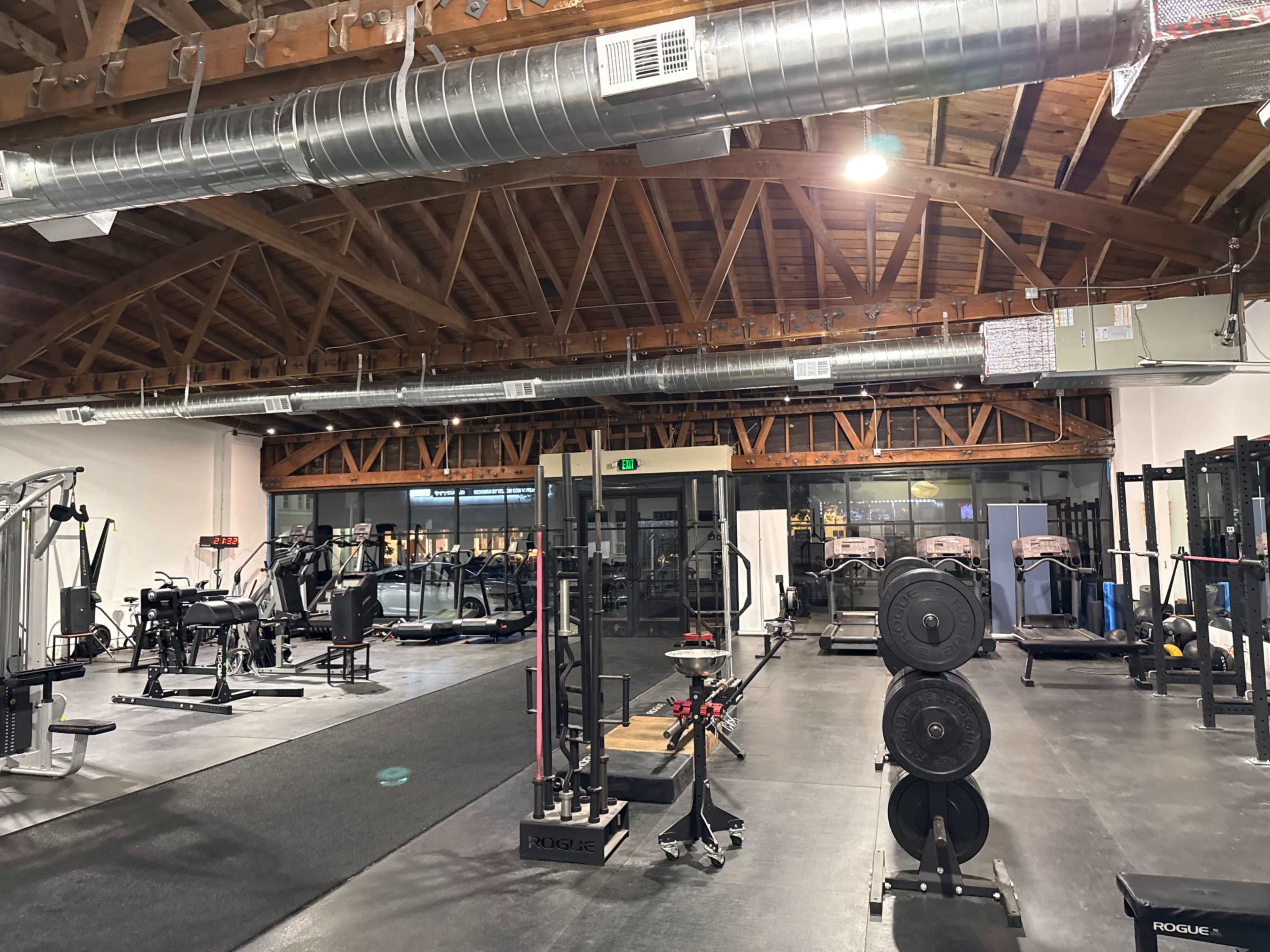 The image shows a spacious gym with various exercise equipment, including weights, machines, and cardio machines, under a wooden beam ceiling.