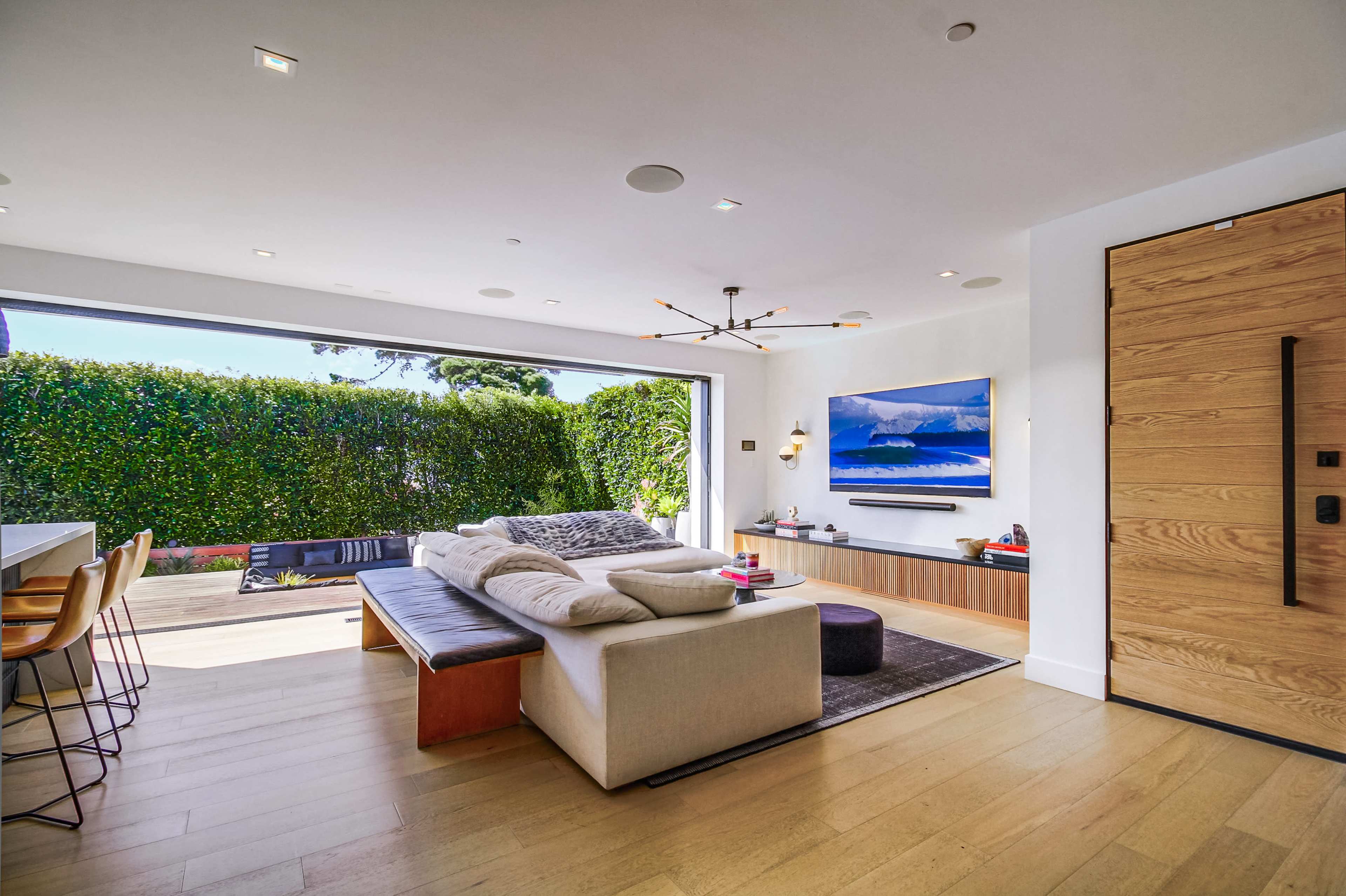The image shows a modern living room with a large sectional sofa, a minimalist entertainment unit, and an open view leading to an outdoor space surrounded by greenery.