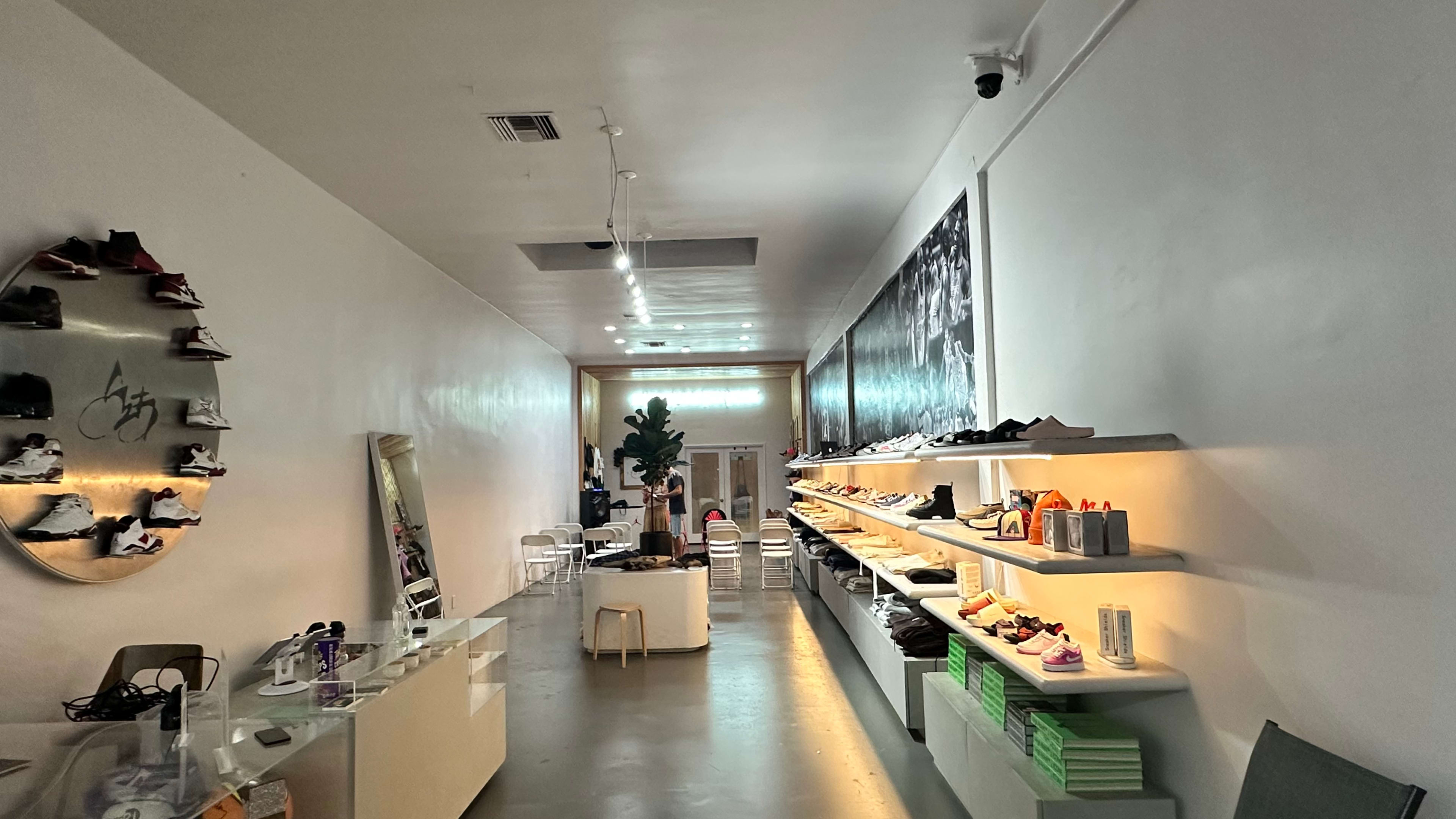The image shows a narrow retail space displaying various sneakers on shelves along the walls, with a mirror and seating area visible.